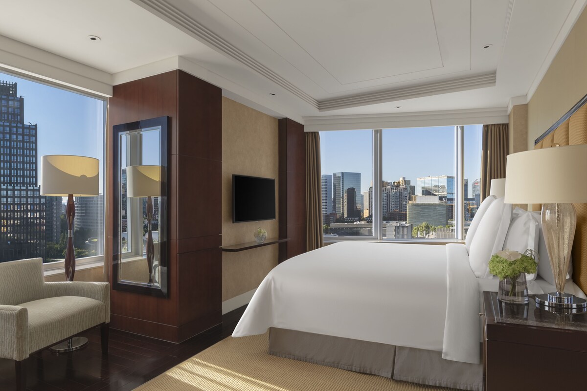 A modern hotel room with a large bed, a TV, a chair, and a view of the city skyline.