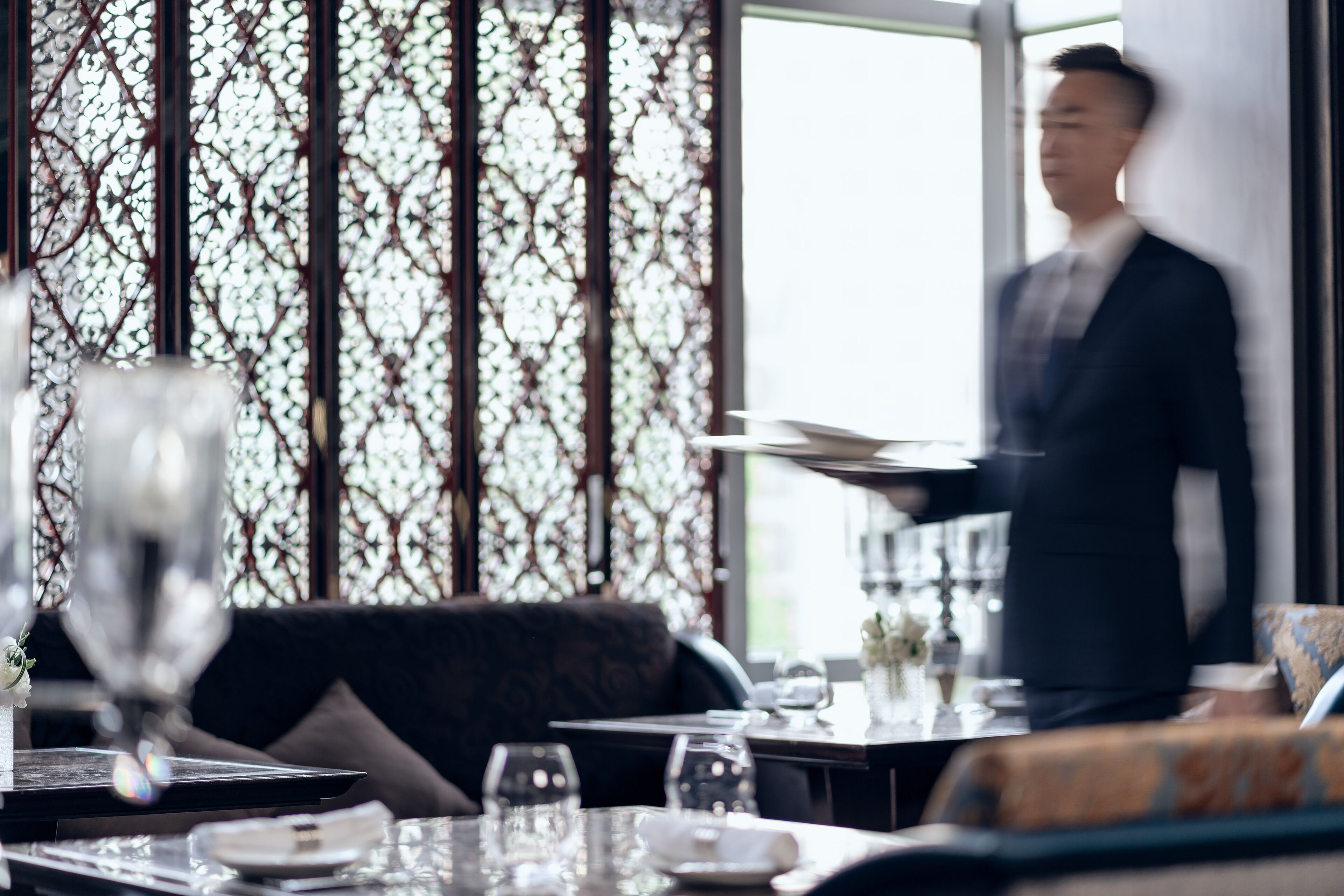 A dining area with a table set for two, a waiter in motion, and a decorative window with a patterned design.