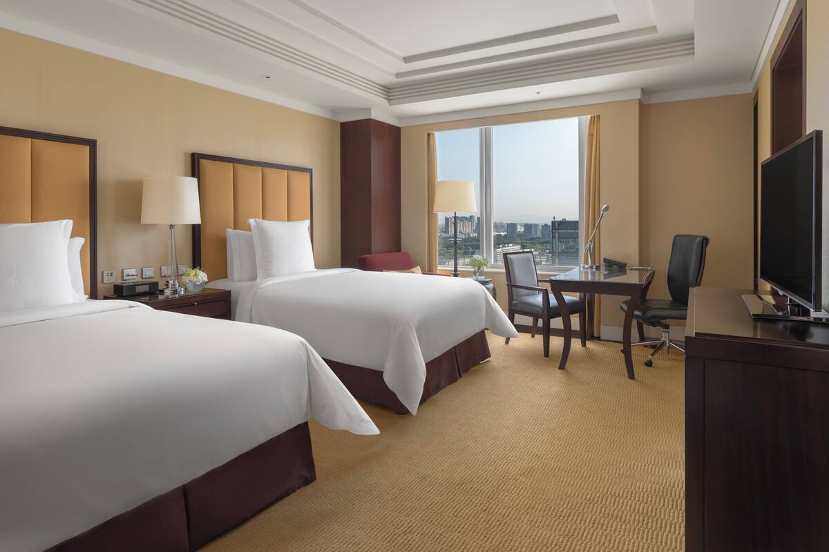 A hotel room with two beds, a desk, a chair, a TV, and a large window offering a city view.