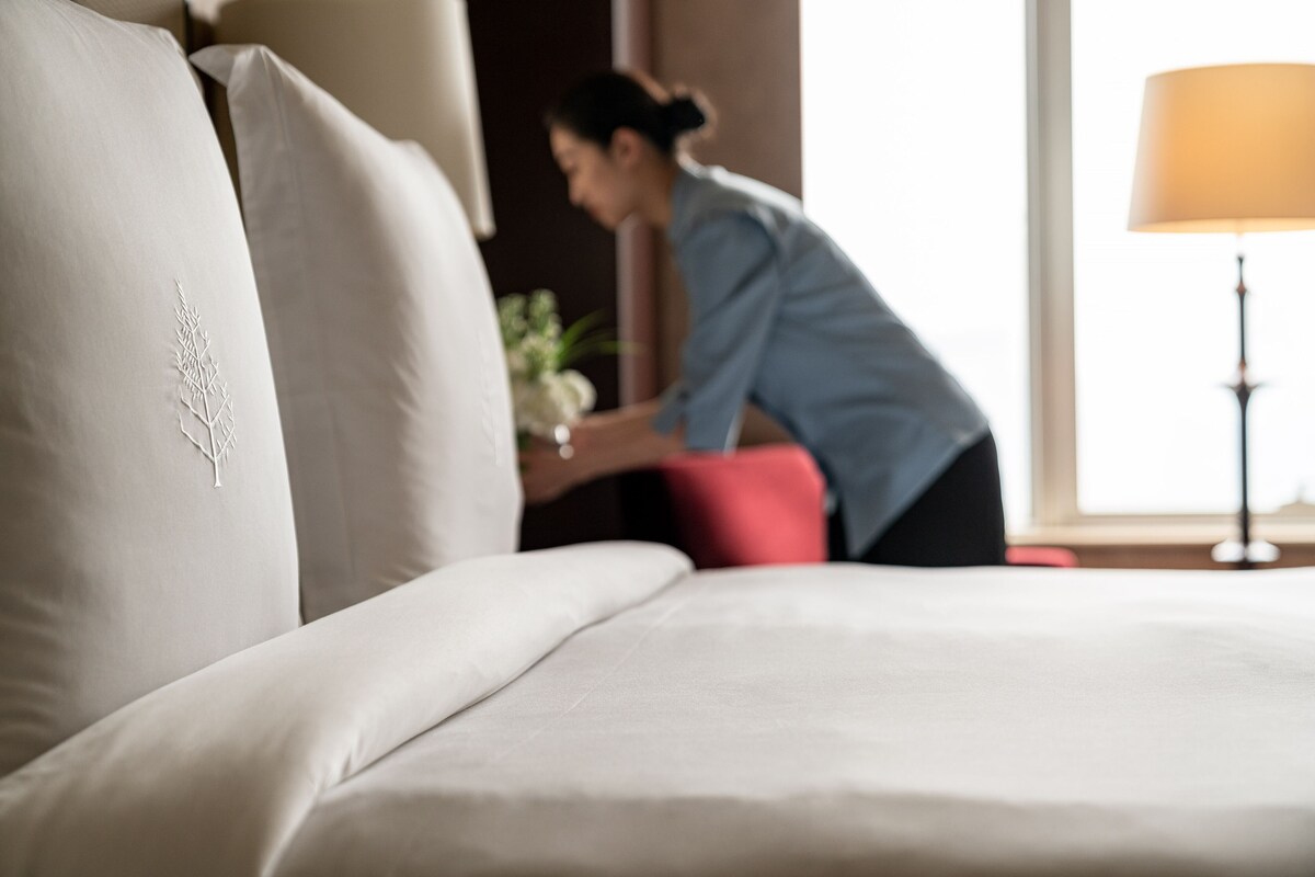 A hotel room with a large bed, a red accent pillow, and a lamp in the corner.