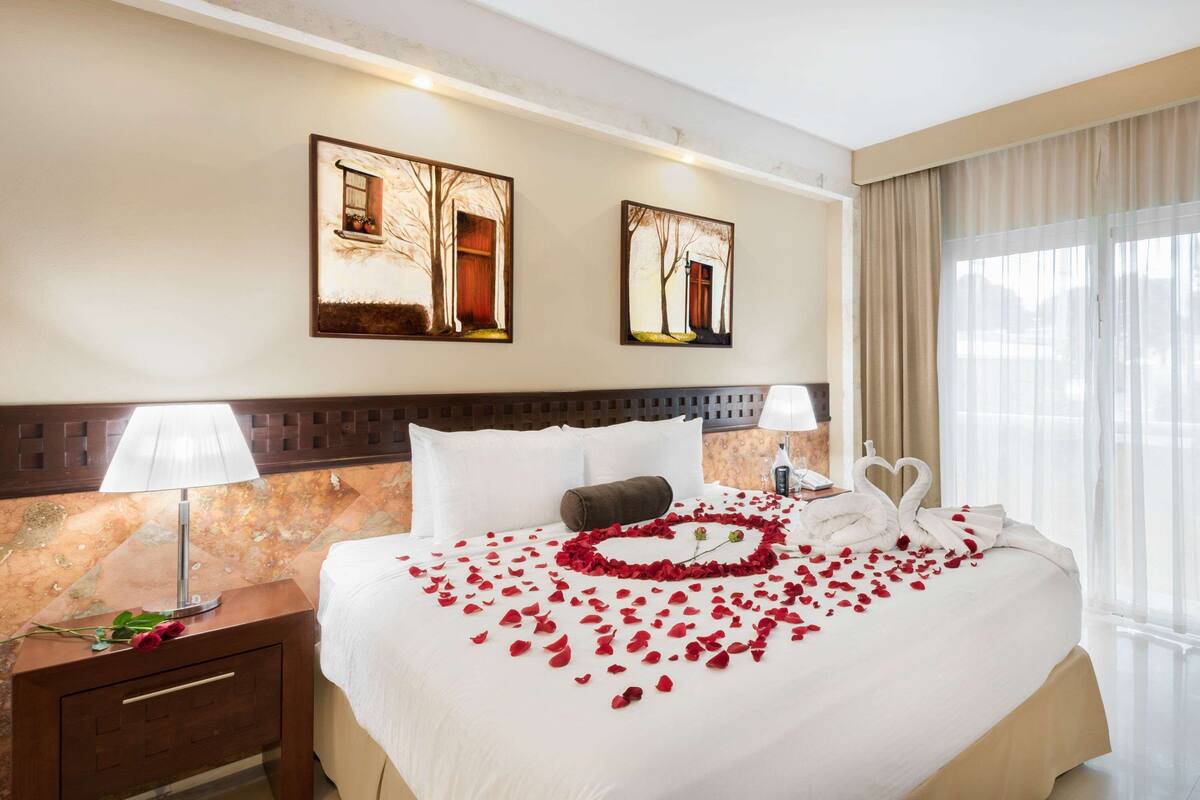 A hotel room with a large bed, two wall-mounted paintings, a nightstand with a lamp, and a window with curtains.