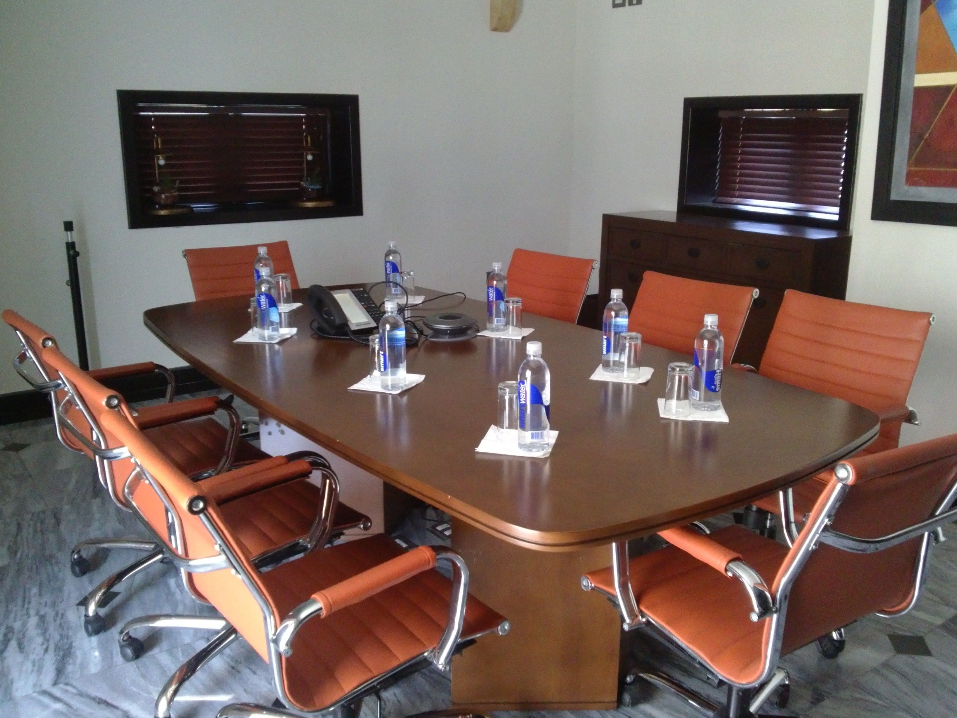 A conference room with a large table, orange chairs, water bottles, a phone, and a window with blinds.