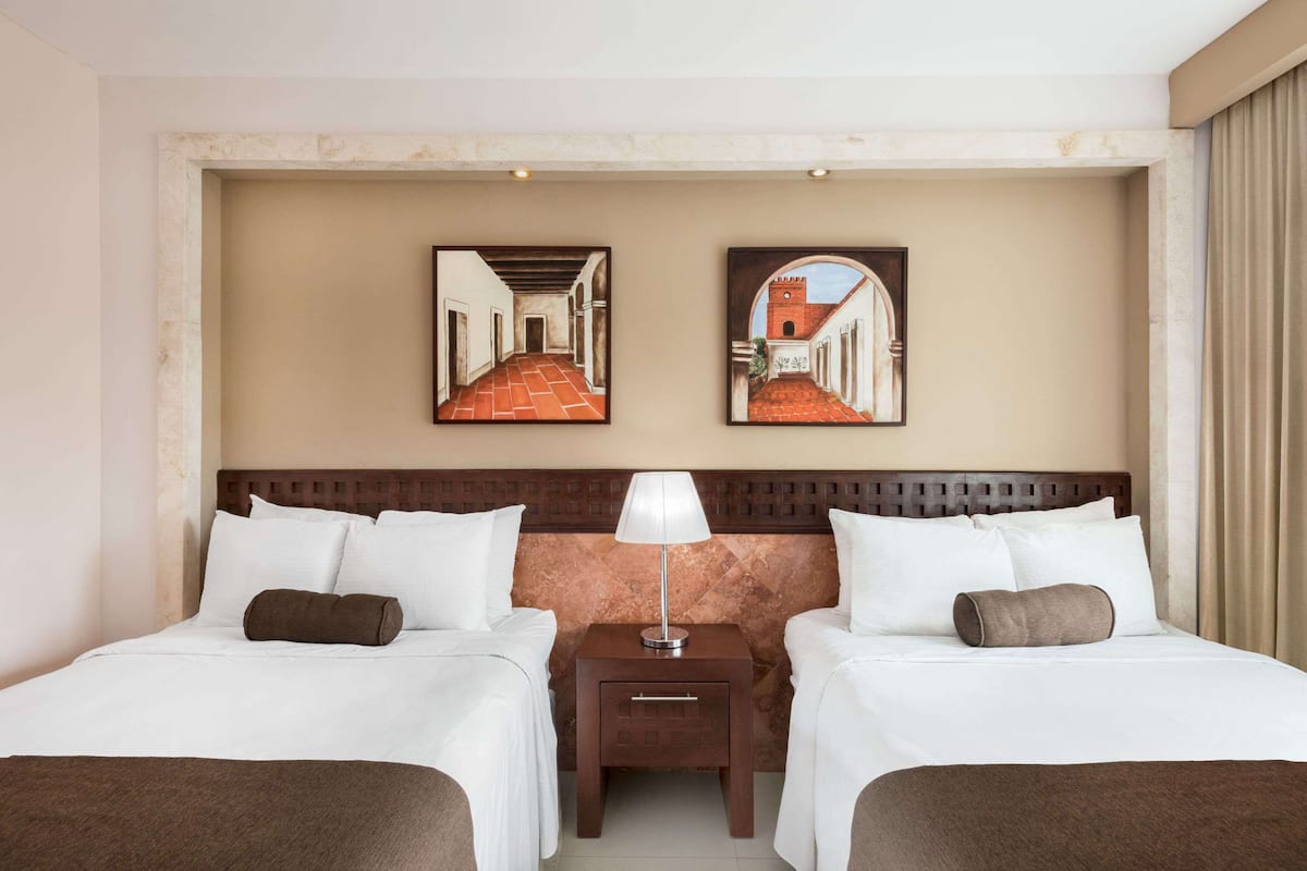 A hotel room with two beds, a nightstand with a lamp, and two framed pictures on the wall.