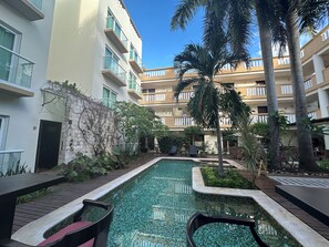 A pool area with tiled flooring, surrounded by a low stone wall, and adjacent to a multi-story building with balconies.