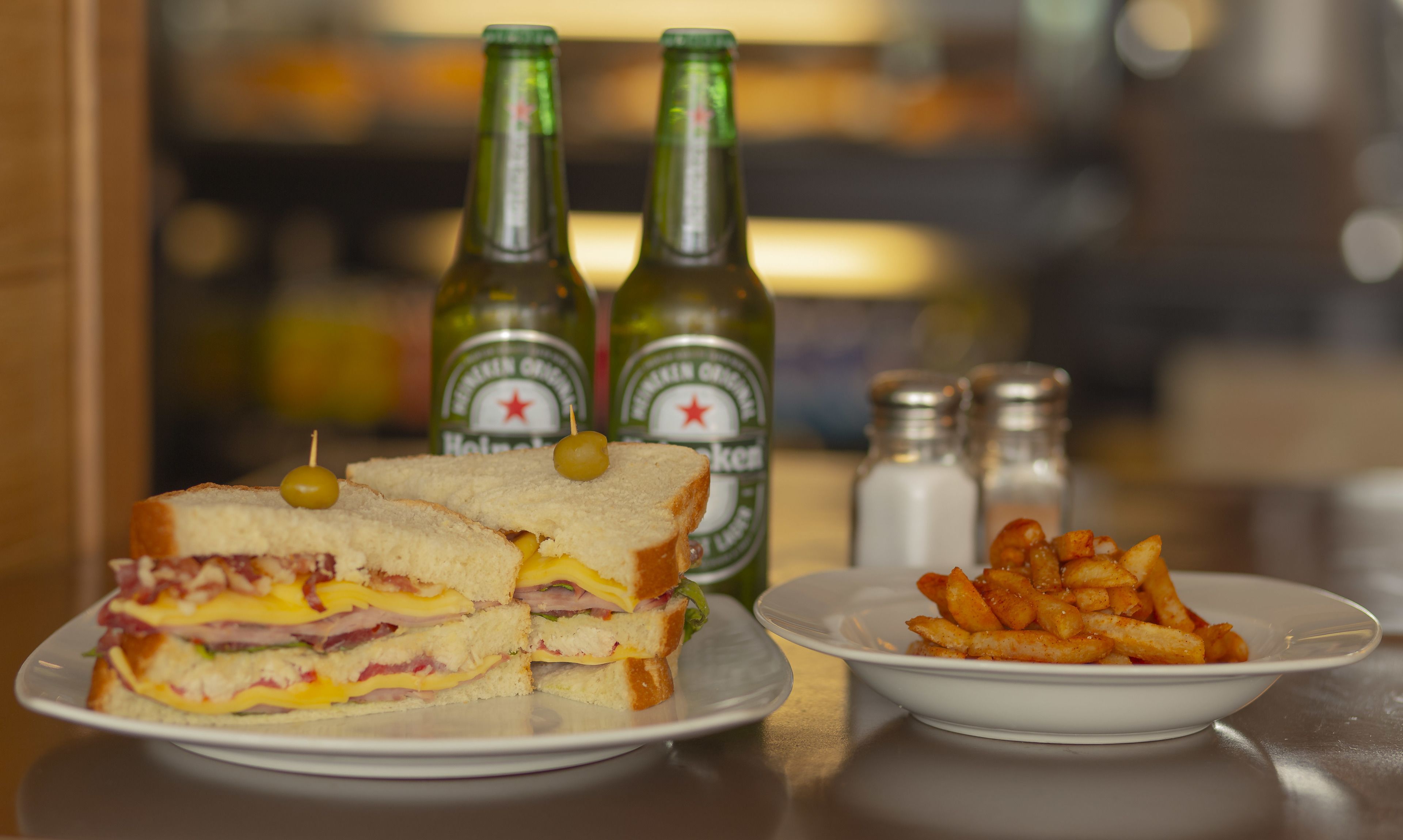 A sandwich with ham, cheese, and a green olive on a plate, accompanied by a bowl of fries and two bottles of beer in the background.