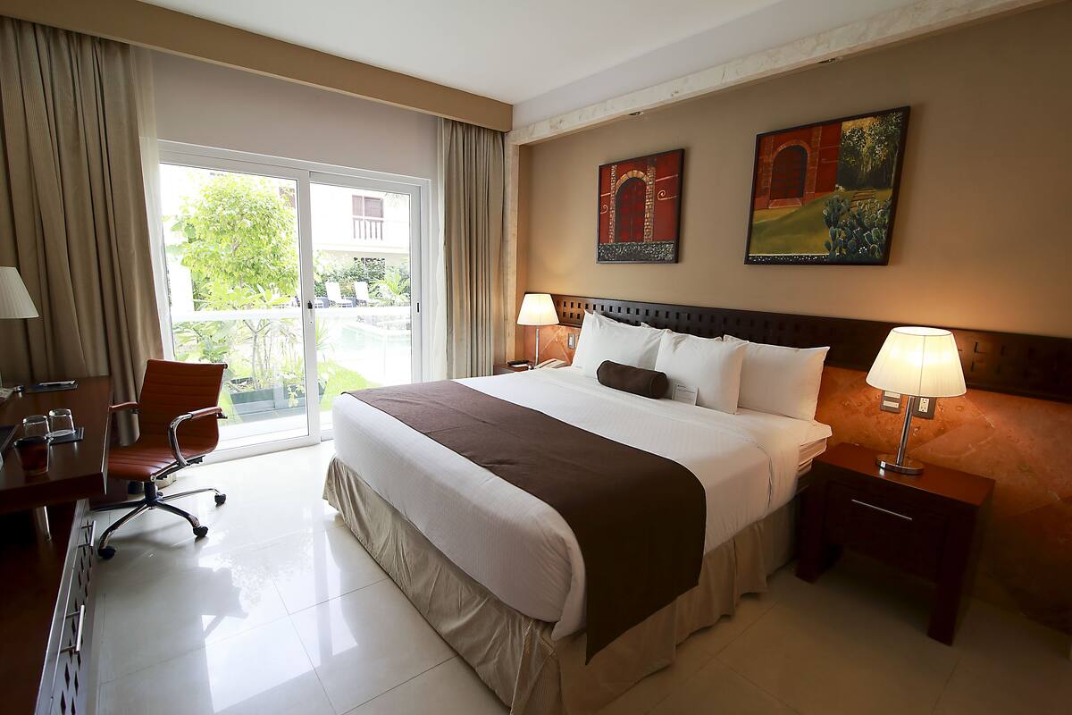 A hotel room with a large bed, two bedside tables with lamps, a desk with a chair, and a view of the outdoors through a sliding door.