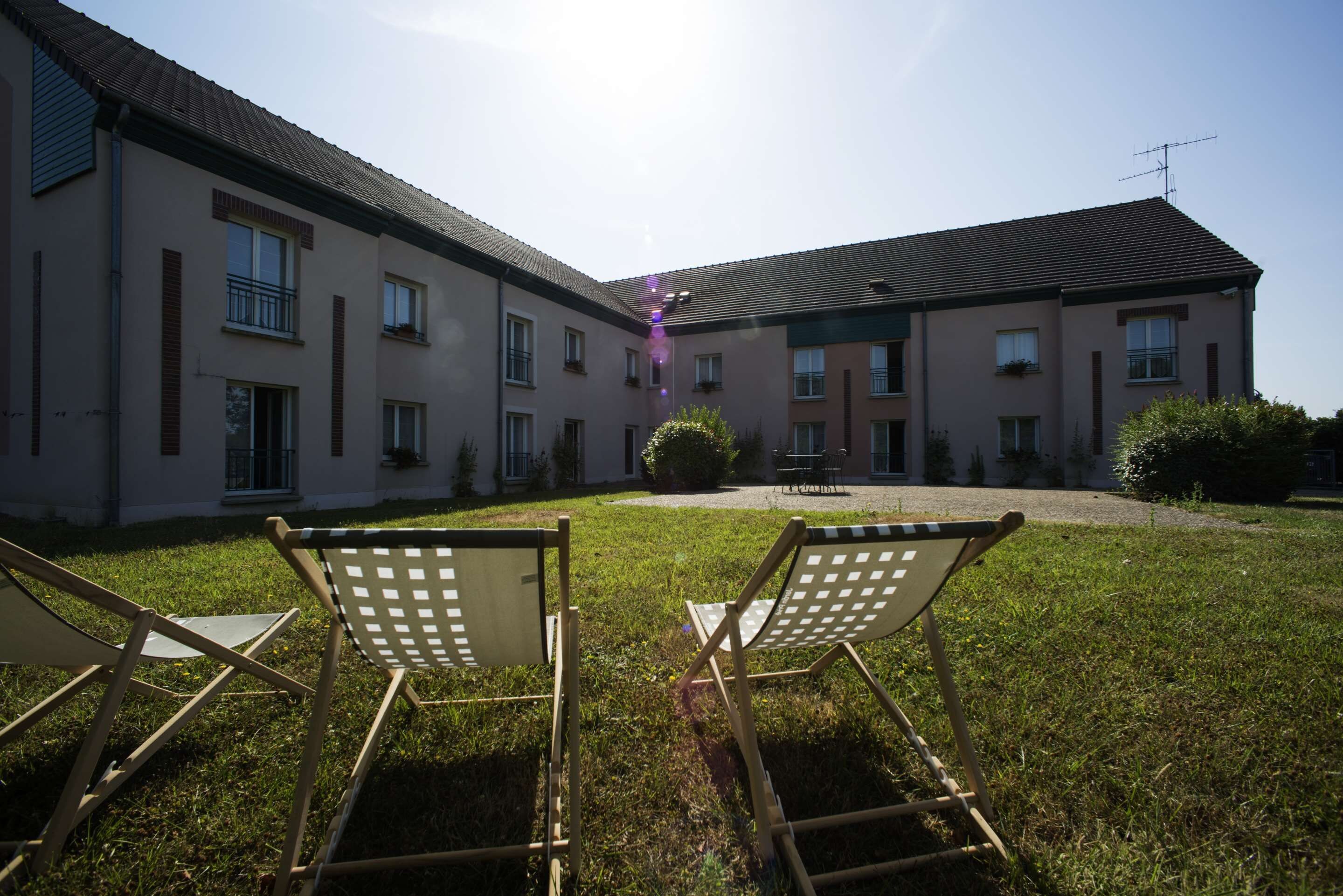 Um pátio com duas cadeiras de descanso, um edifício com várias janelas e sacadas, e um céu claro.