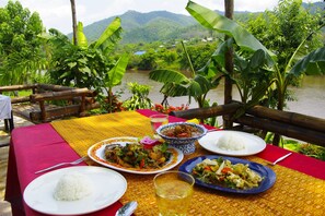 A table set with various dishes, including rice, a bowl of meat, and a plate of vegetables, overlooking a river with lush greenery and hills in the background. - Areeya Phuree Resort (Mae Ai)