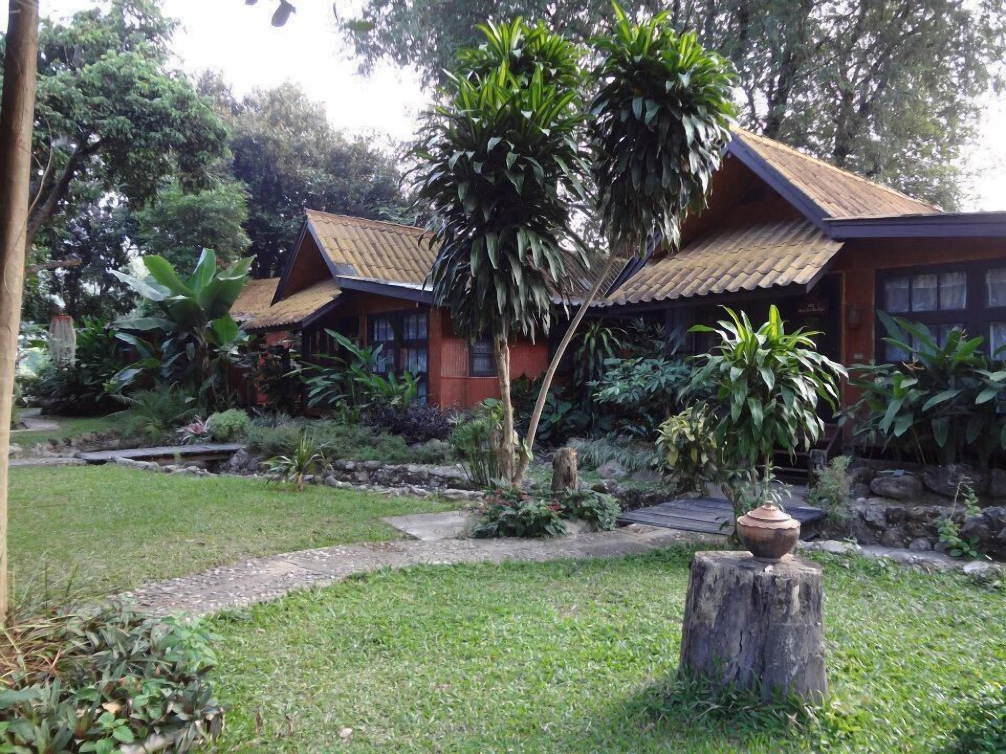 A traditional wooden house with a tiled roof, surrounded by lush greenery and a stone pathway.