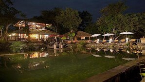 A nighttime view of a resort with a pool, thatched-roof structures, and umbrellas. - Zen Namkhan Boutique Resort (Luang Prabang)