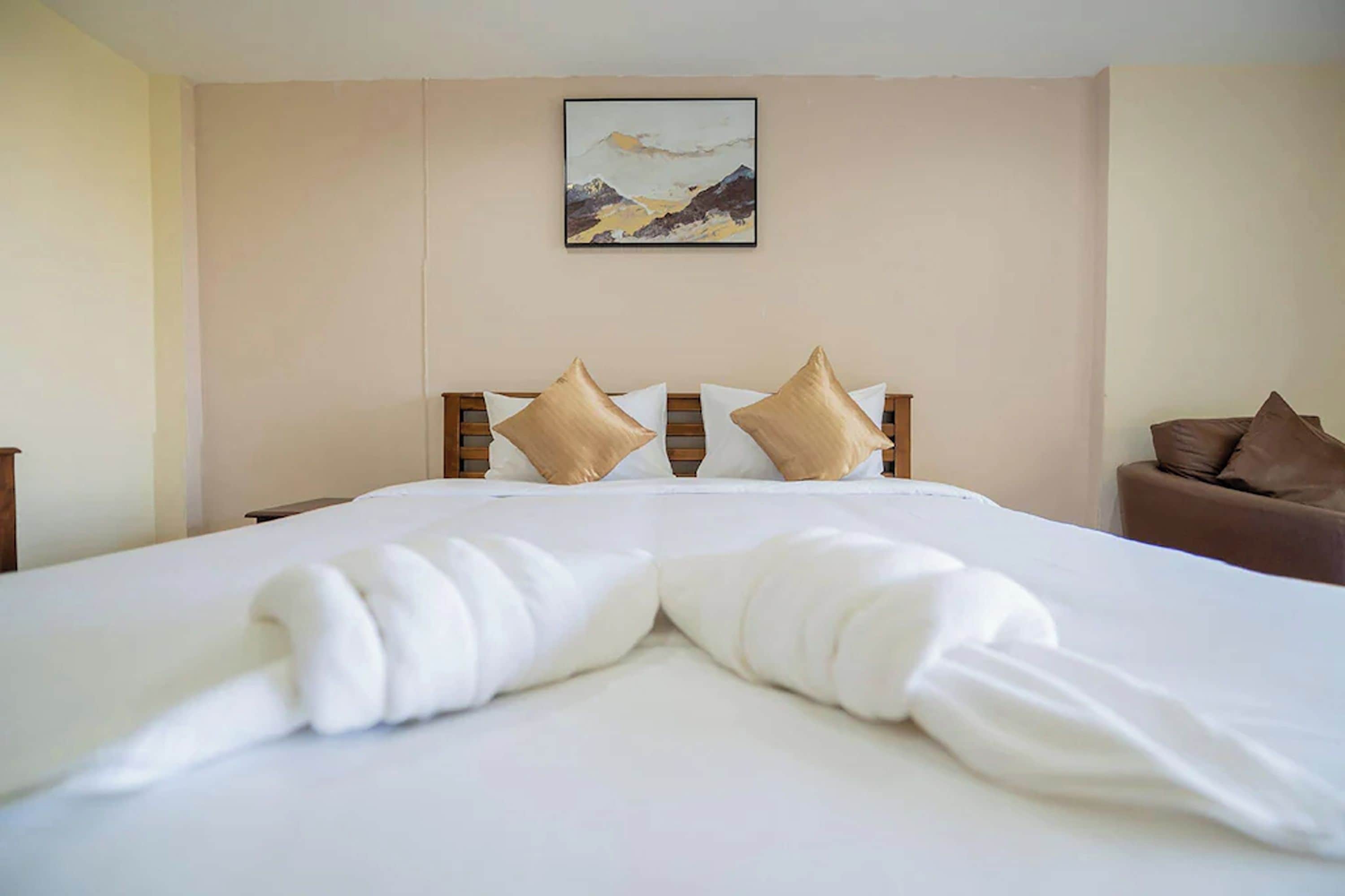 A hotel room with a neatly made bed, a painting on the wall, and a brown armchair.