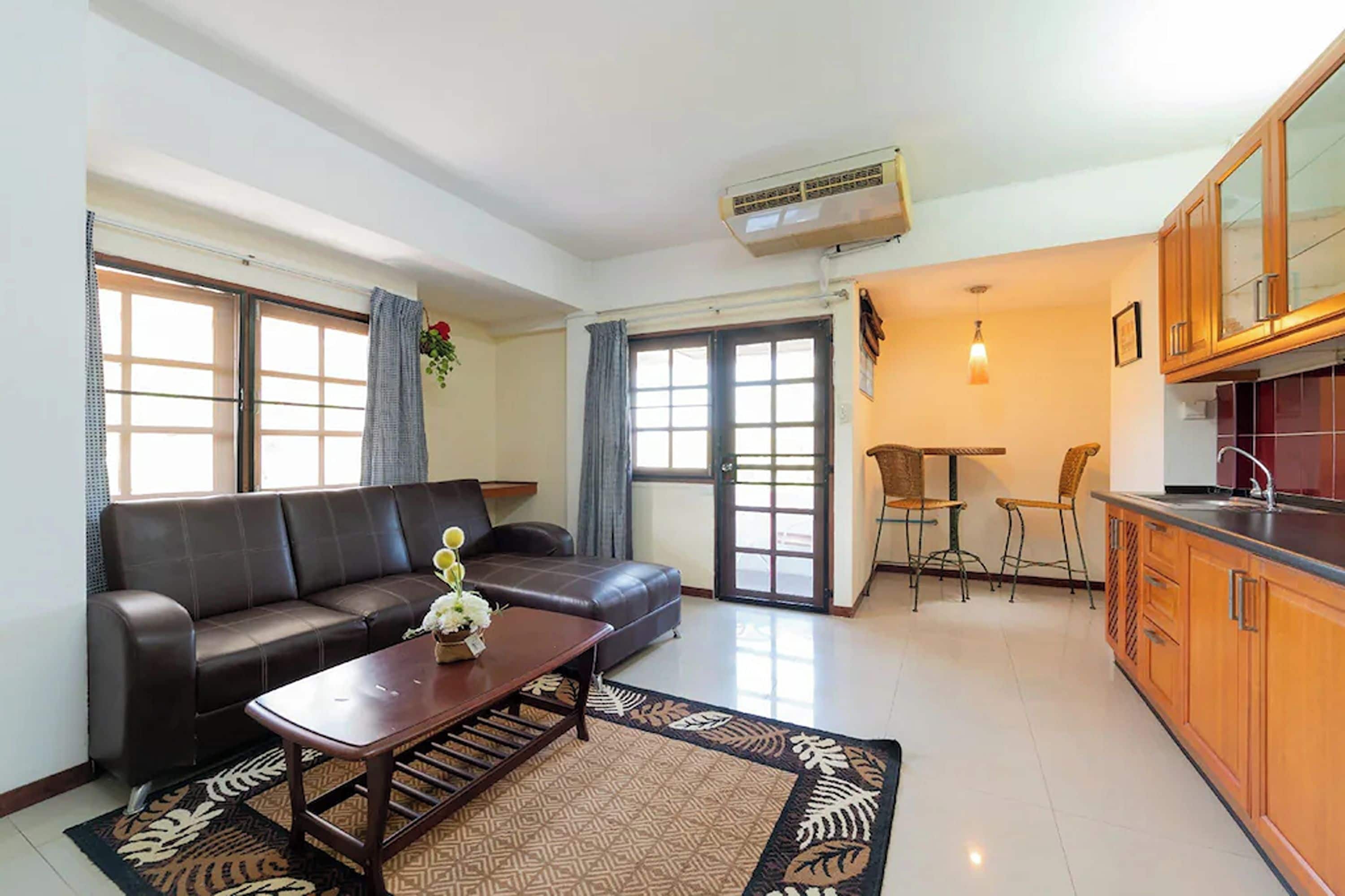 A living room with a sofa, coffee table, and a rug. There is a kitchen area with wooden cabinets and a dining table with chairs.