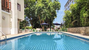 A swimming pool with tiled edges, surrounded by lounge chairs and umbrellas, with trees and a building in the background. - Hillside Resort Pattaya (Pattaya)