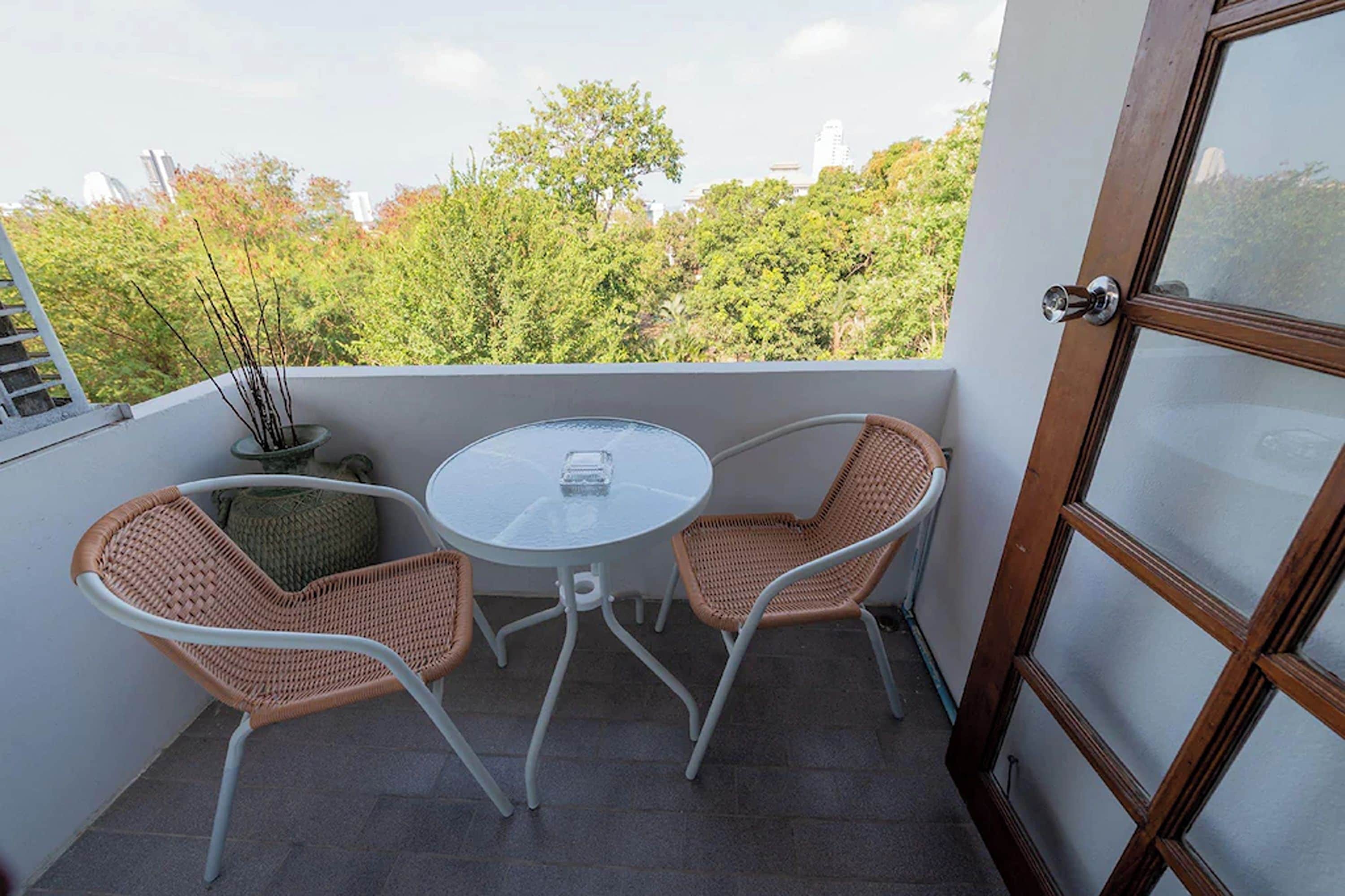 En balkon med et rundt bord, to fletstole, en potteplante og udsigt til træer og bygninger.