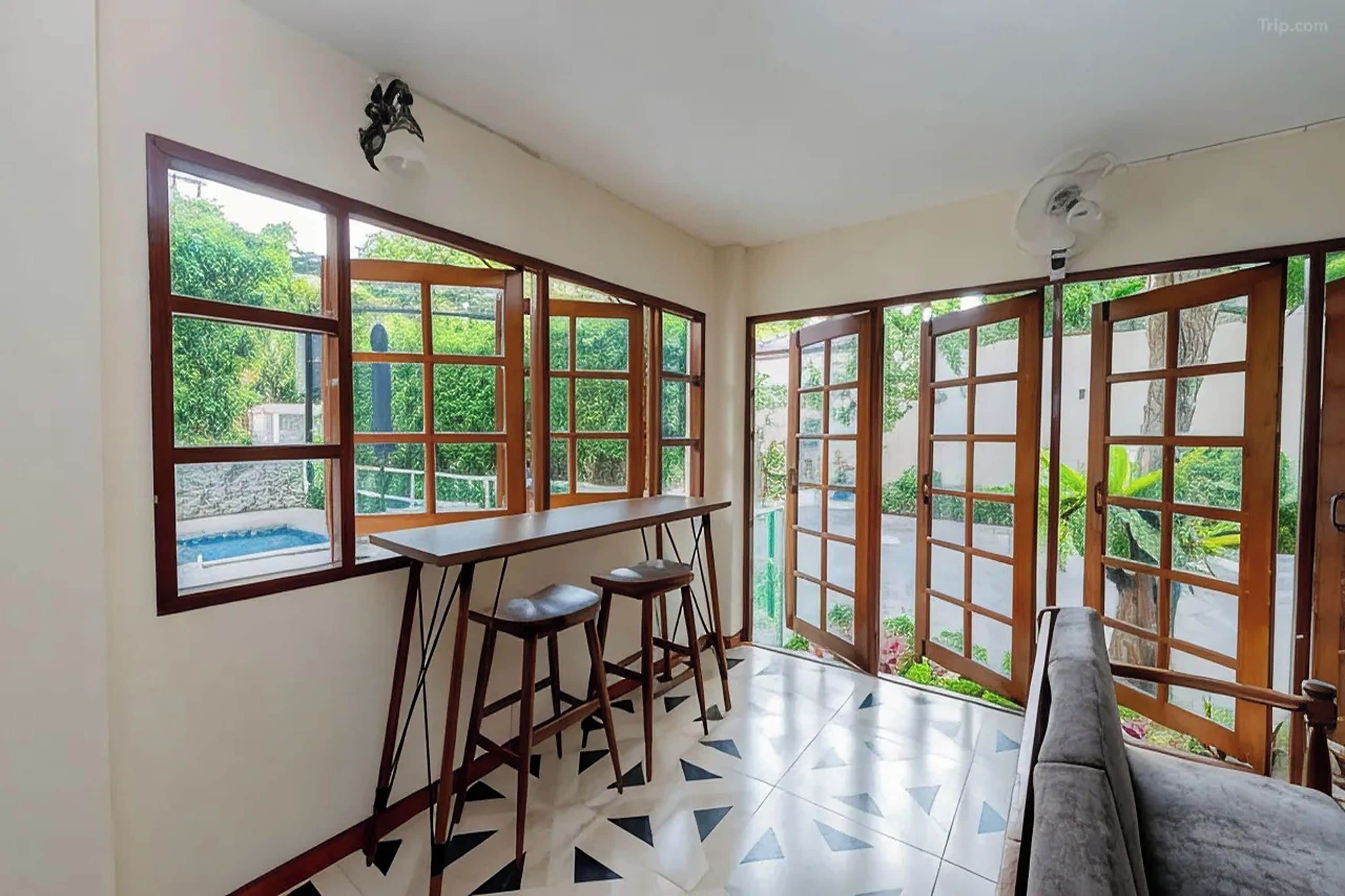 A room with a bar area, a sofa, and large windows with a view of greenery.
