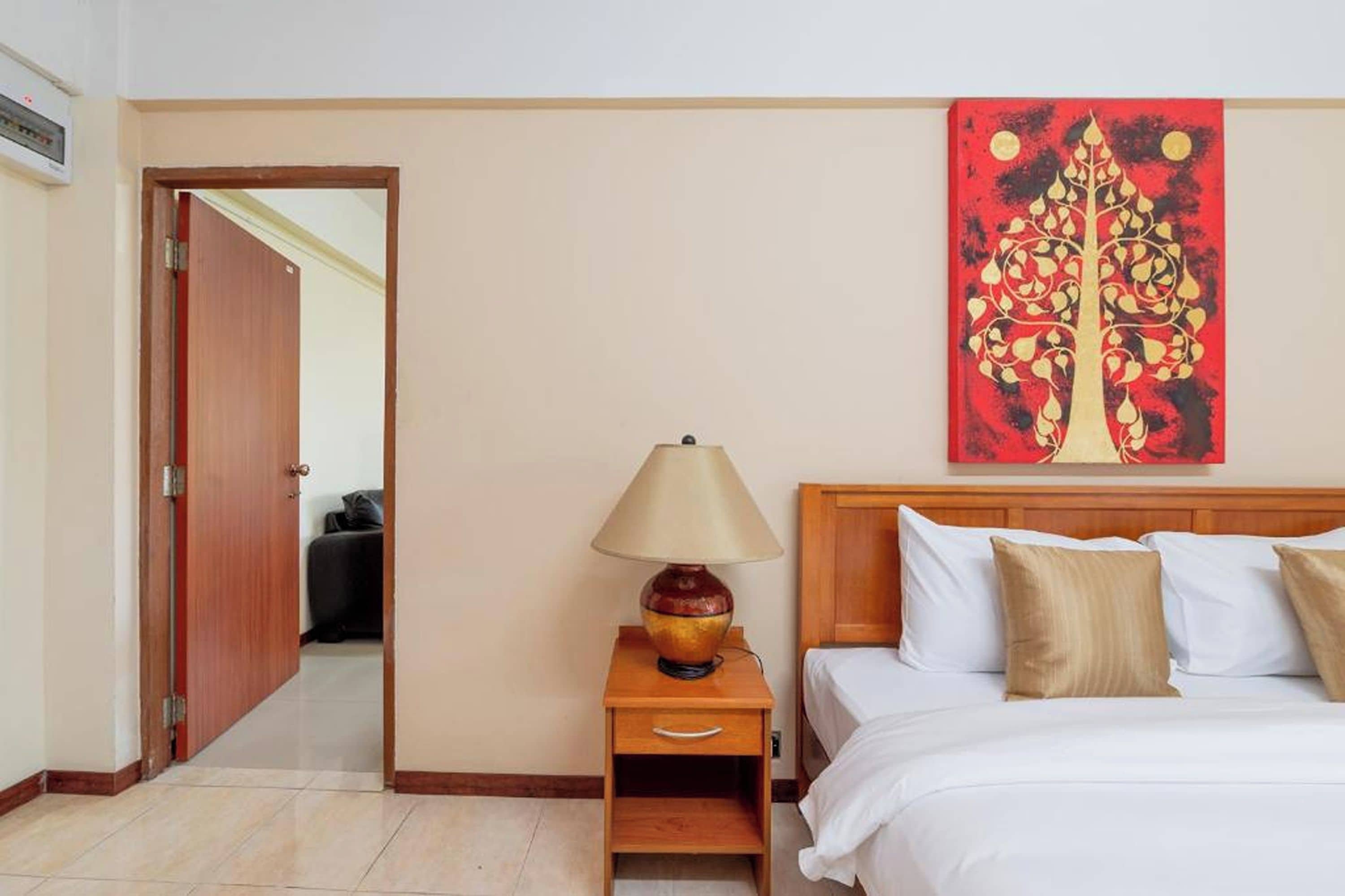 A hotel room with a bed, a nightstand, a lamp, a painting, and a door leading to another room.