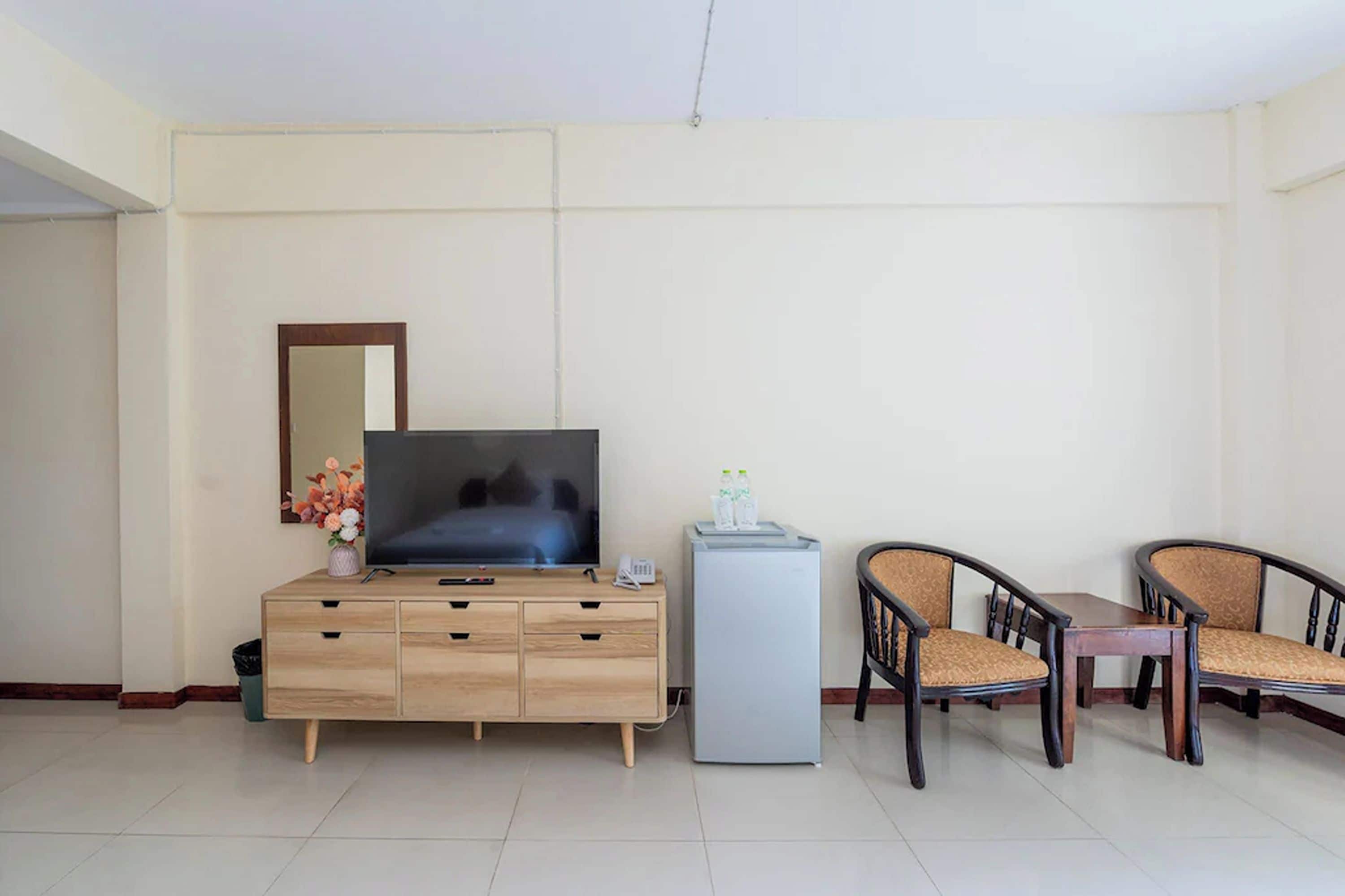 A hotel room with a wooden cabinet, a flat-screen TV, a small refrigerator, two wicker chairs, and a mirror on the wall.