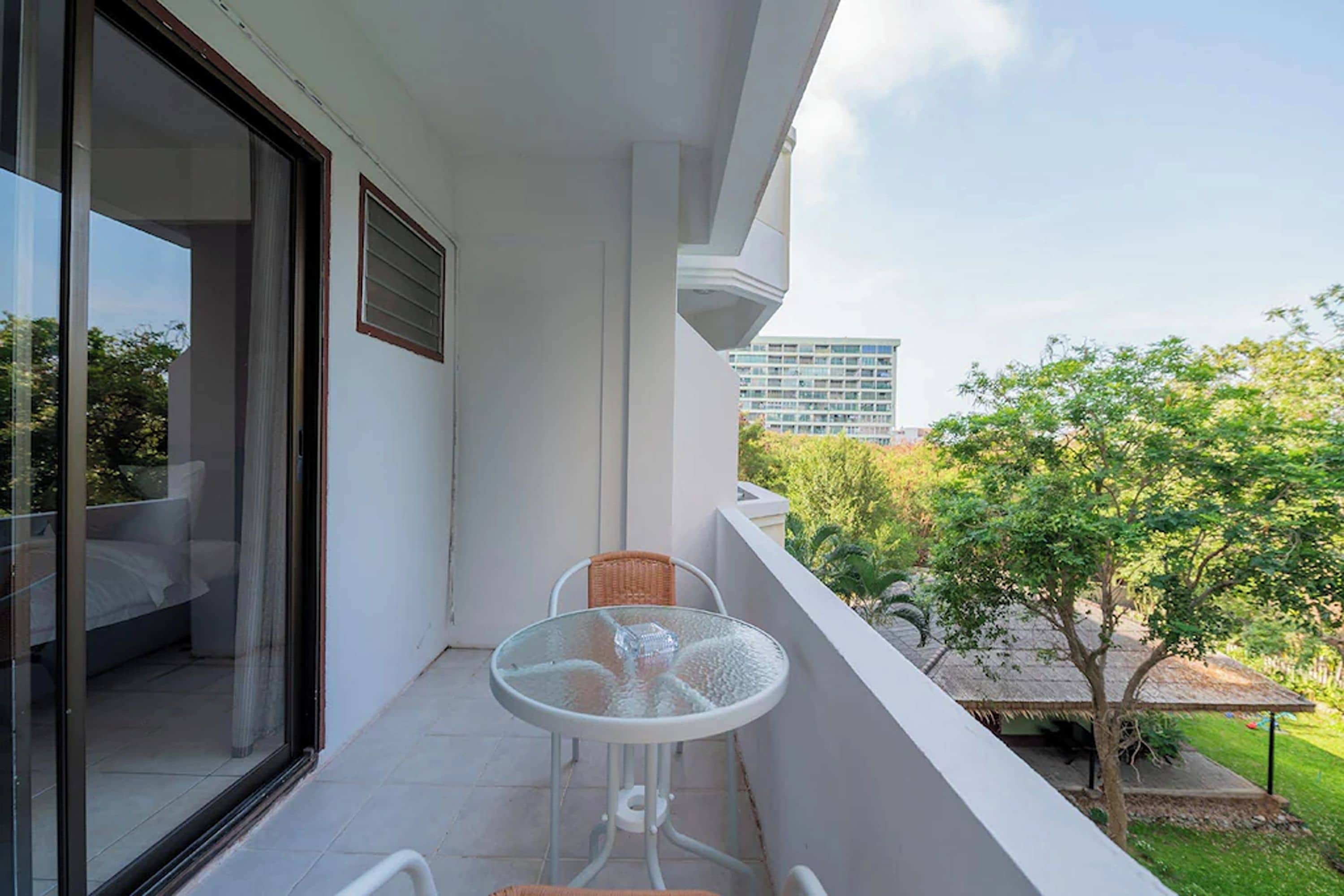 A balcony with a glass door, a small table with a chair, and a view of a building and trees.