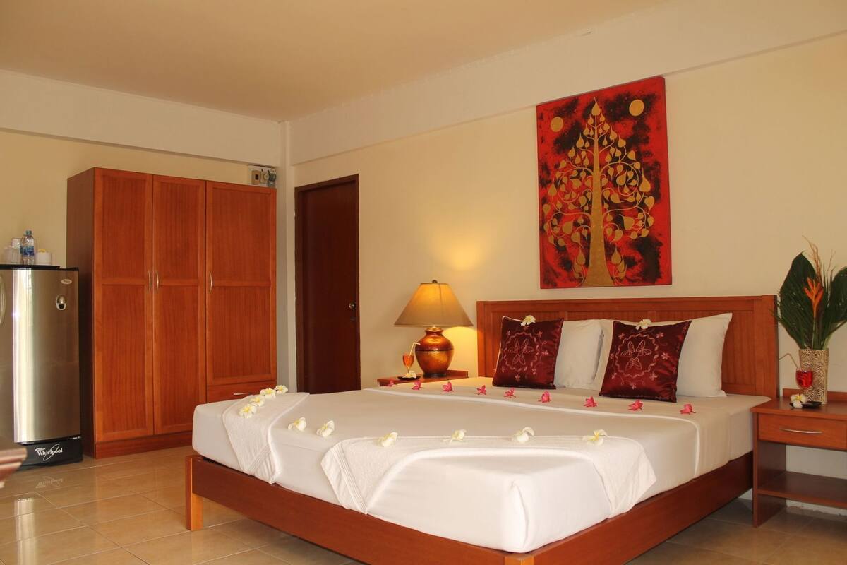 A hotel room with a bed, a wooden wardrobe, a refrigerator, a lamp, and a painting on the wall.