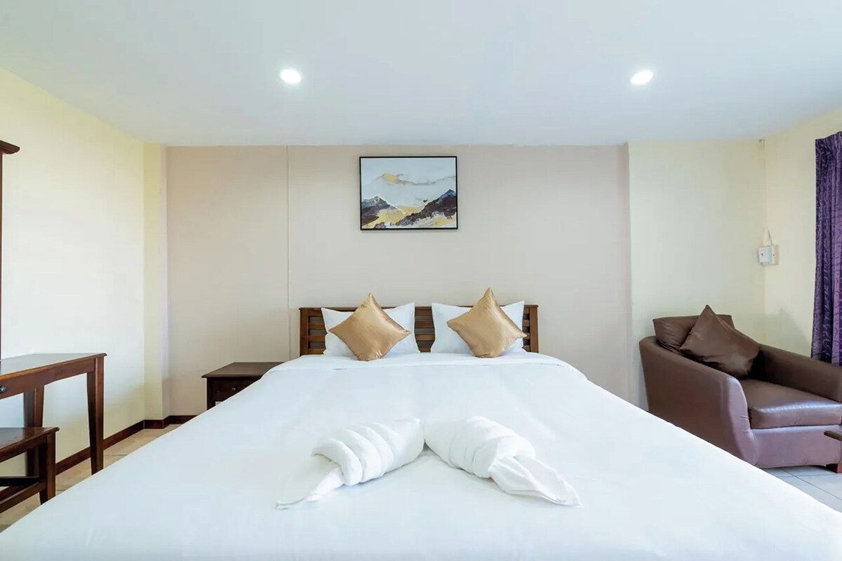 A hotel room with a large bed, a wooden headboard, a painting on the wall, a brown leather armchair, and a small table.