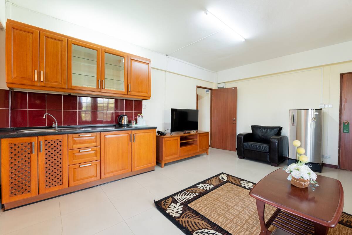 A modern kitchen with wooden cabinets, a black countertop, and a built-in TV. There is a black sofa and a refrigerator in the living area.