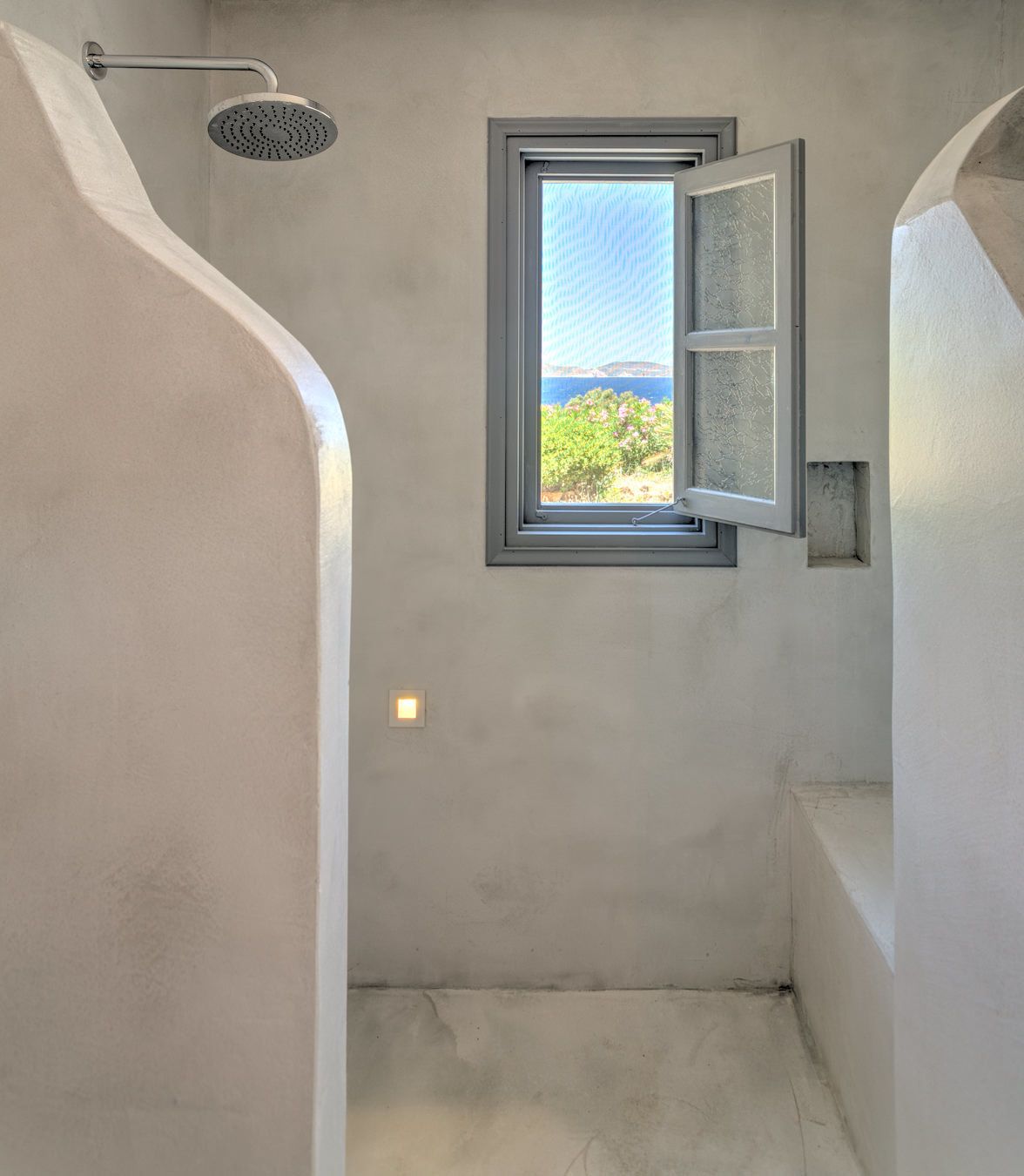 A bathroom with a shower, a window with a view of greenery and a body of water, and a light fixture.