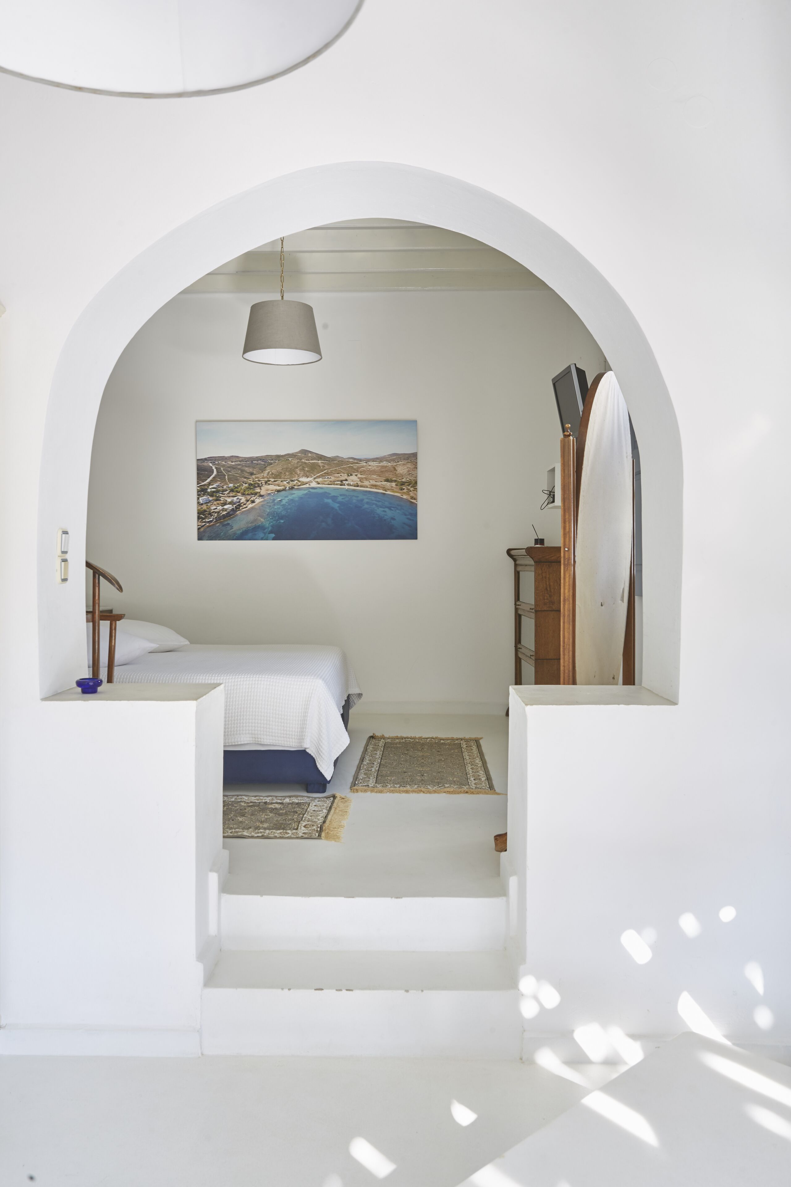 Un dormitorio con una cama, un cuadro de un paisaje costero, una tabla de surf y un escritorio con una lámpara.