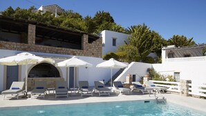 A pool area with lounge chairs and umbrellas, a stone wall, and a white building with a staircase.