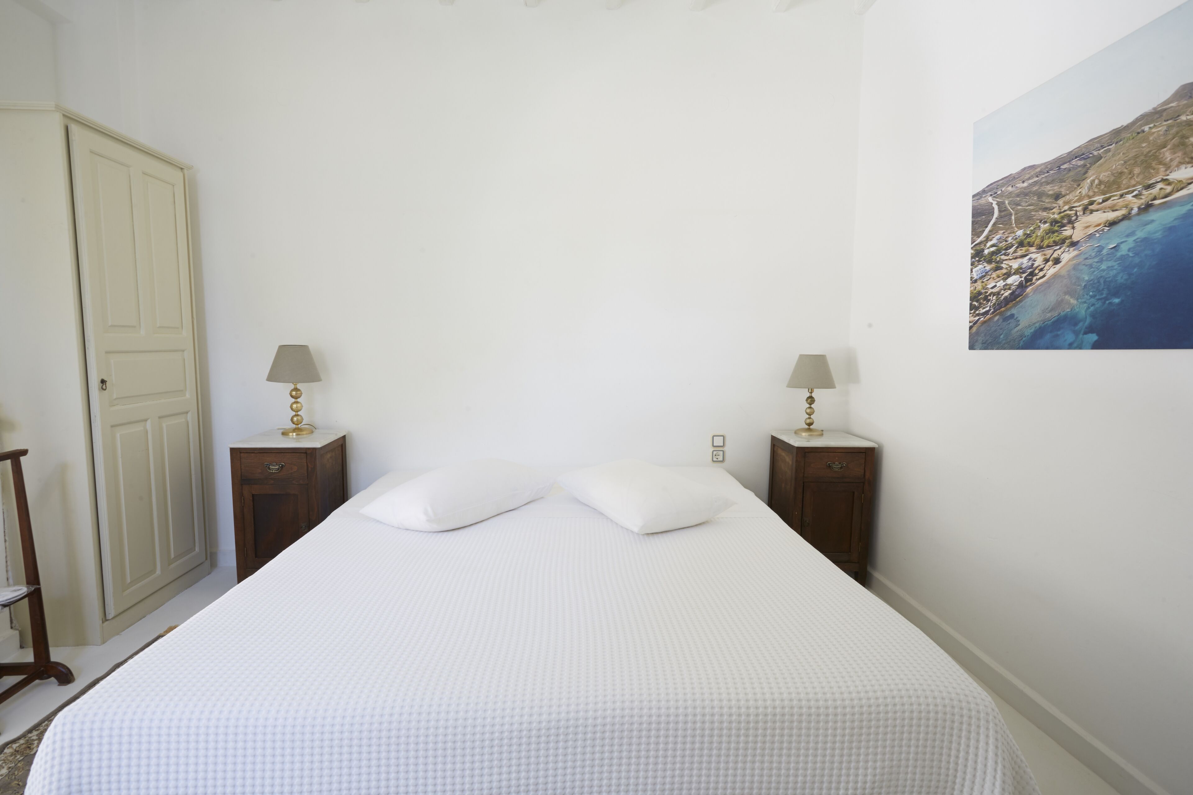 Un dormitorio con una cama, dos mesitas de noche con lámparas, una puerta y un cuadro colgado en la pared.