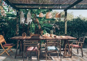 Una mesa de madera al aire libre con sillas, rodeada de abundante vegetación y cestas colgantes.