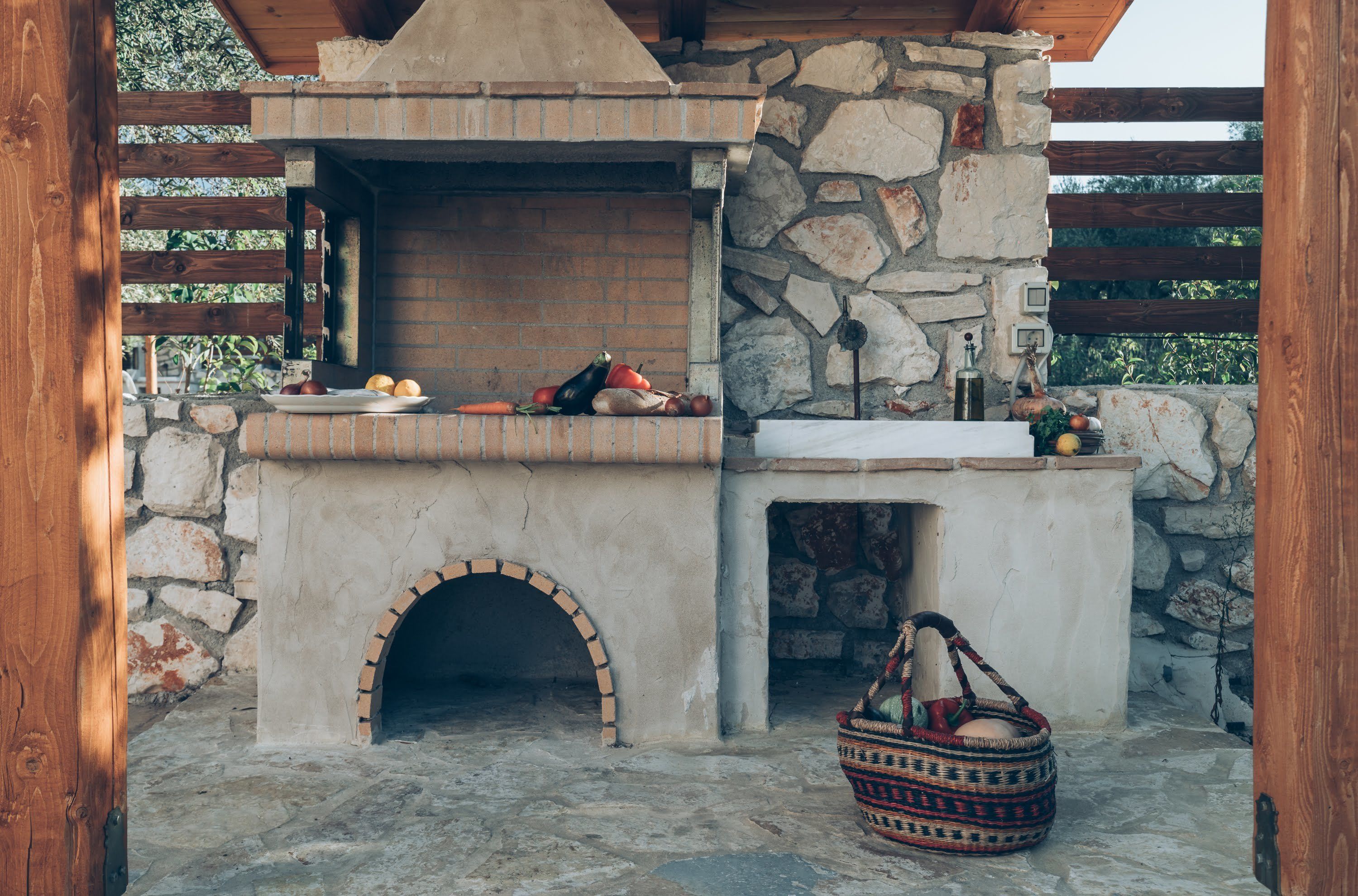 An outdoor brick oven with a stone wall and wooden beams, a basket, and a variety of vegetables on the counter.
