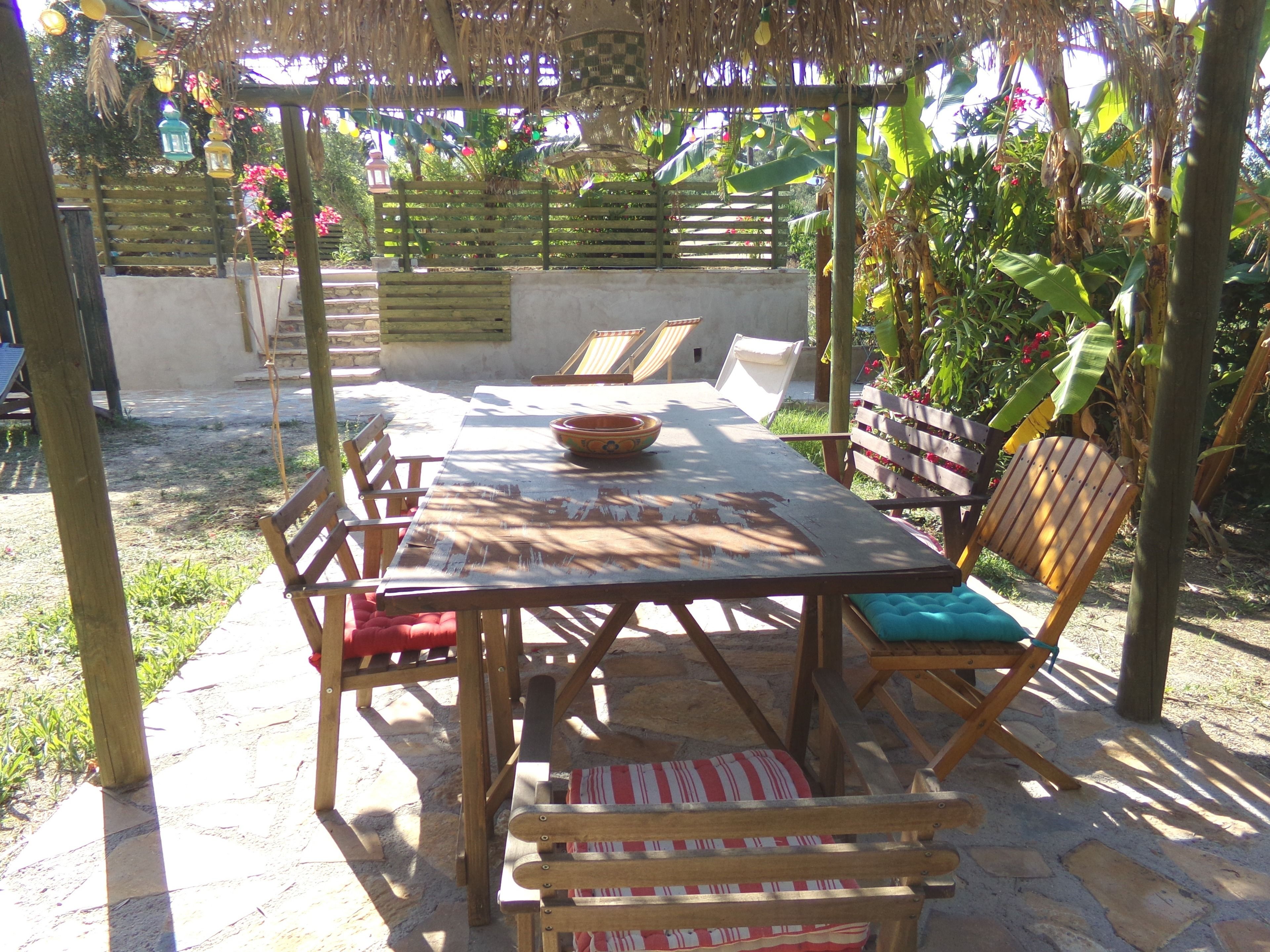 Une terrasse extérieure avec une table et des chaises en bois, entourée de plantes et d’une clôture en bois.