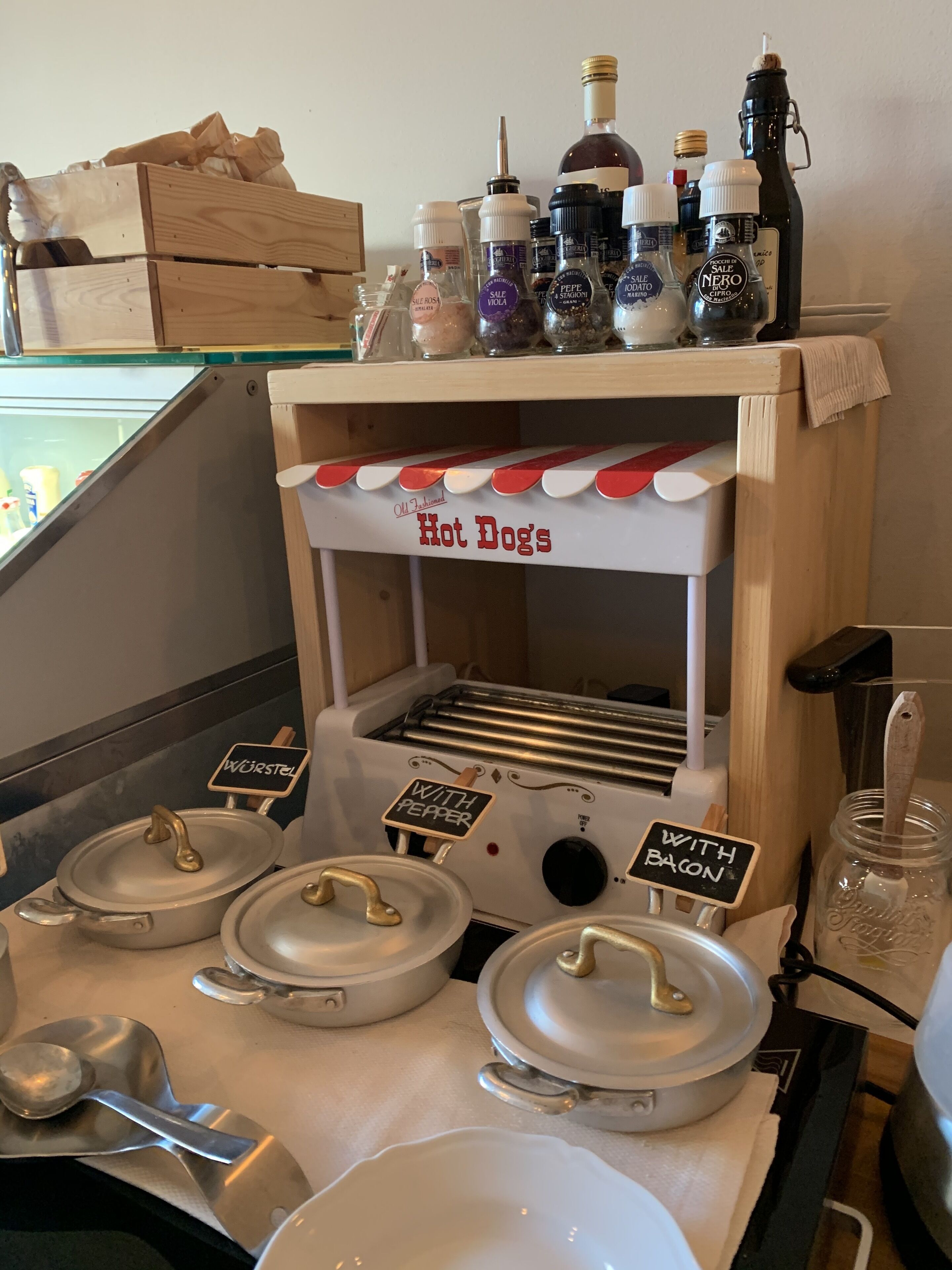 Un coin cuisine avec un comptoir en bois où sont disposés divers condiments et un four à grille-pain sur lequel est écrit « Hot Dogs », avec les options « Wurstel » et « avec du poivre ».