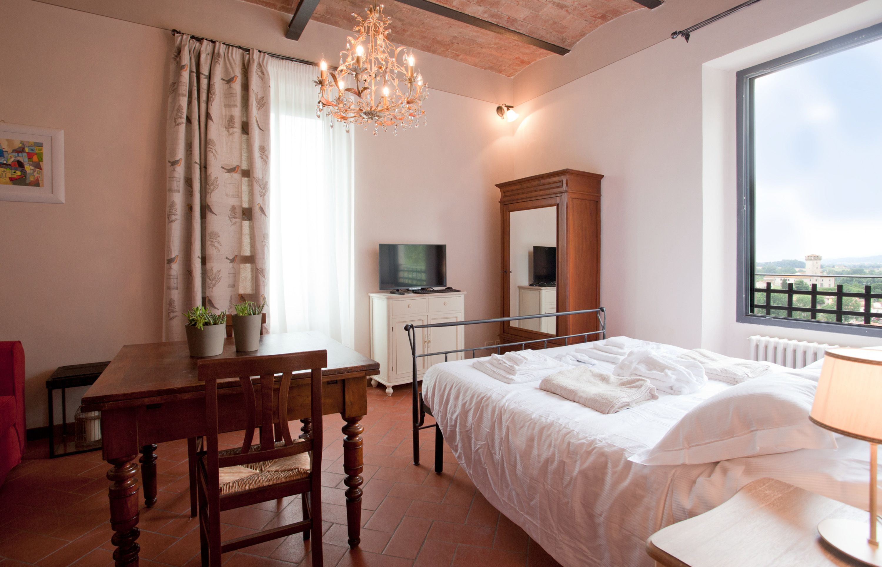 A bedroom with a bed, a desk, a chair, a television, a mirror, and a window with a view.