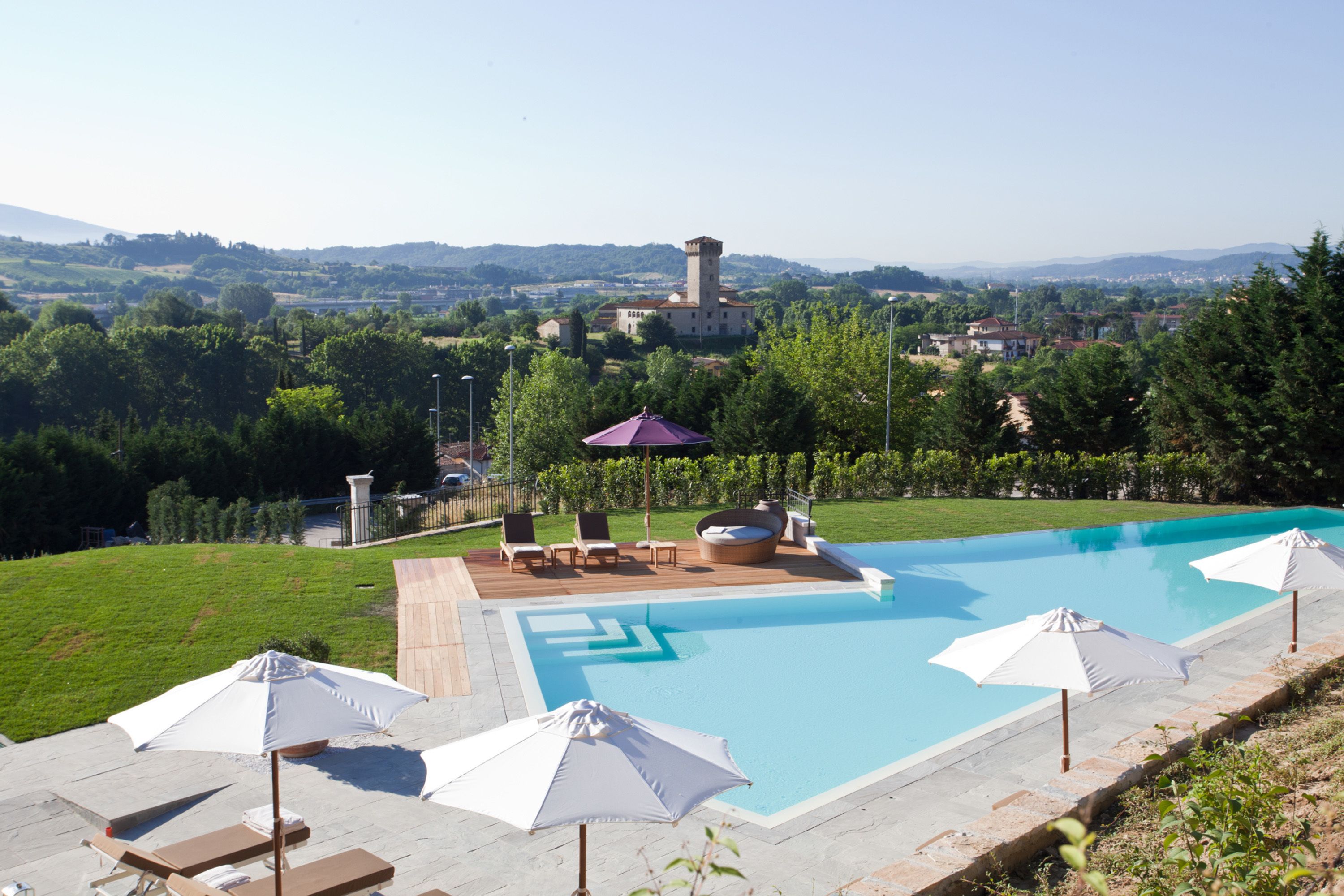 Una piscina con sillas de descanso y sombrillas, rodeada de césped y con vistas a un castillo a lo lejos.