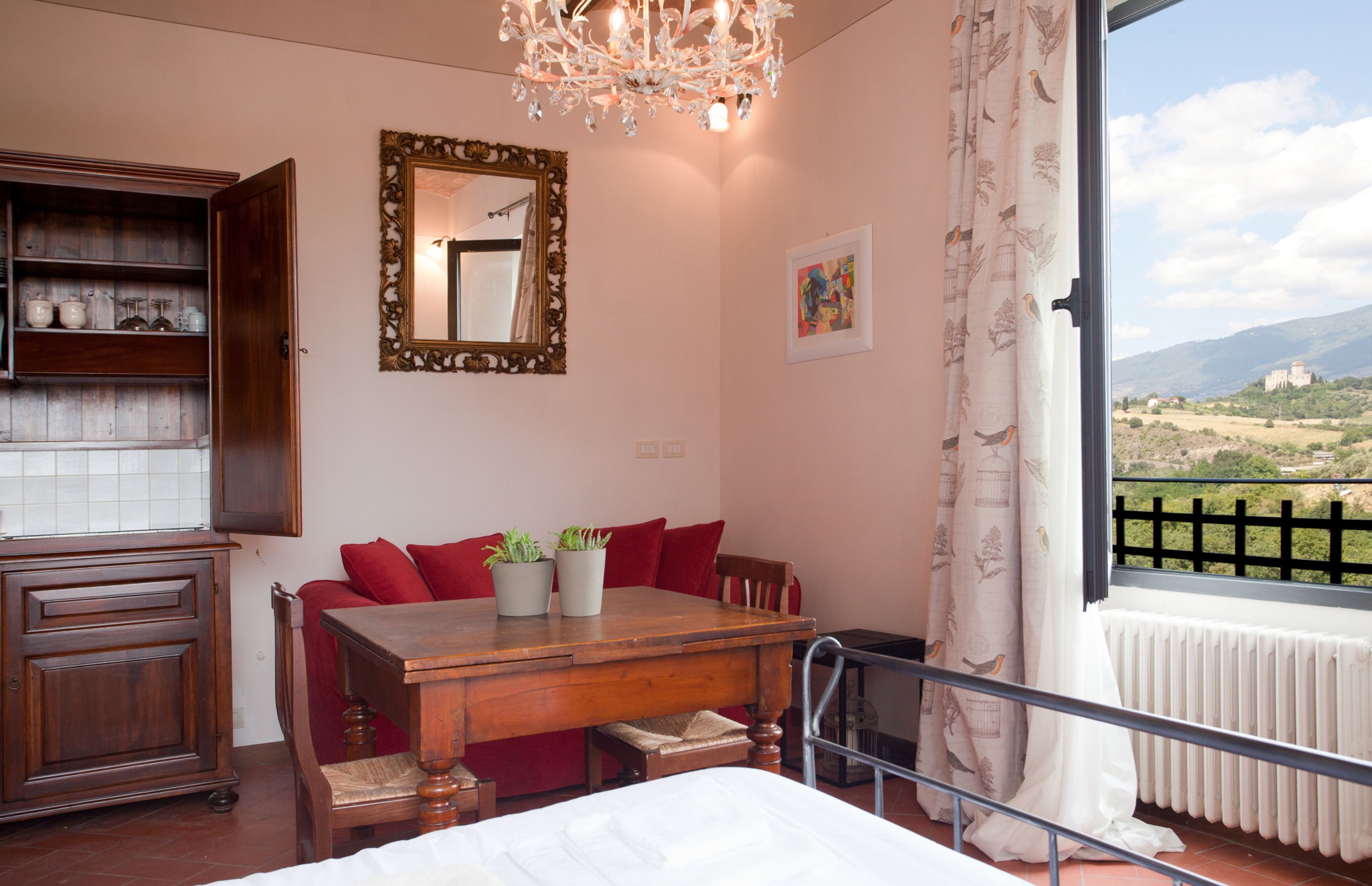 Quarto com cama de madeira, mesa de jantar com almofadas vermelhas, um candelabro, um espelho e vista para o campo.