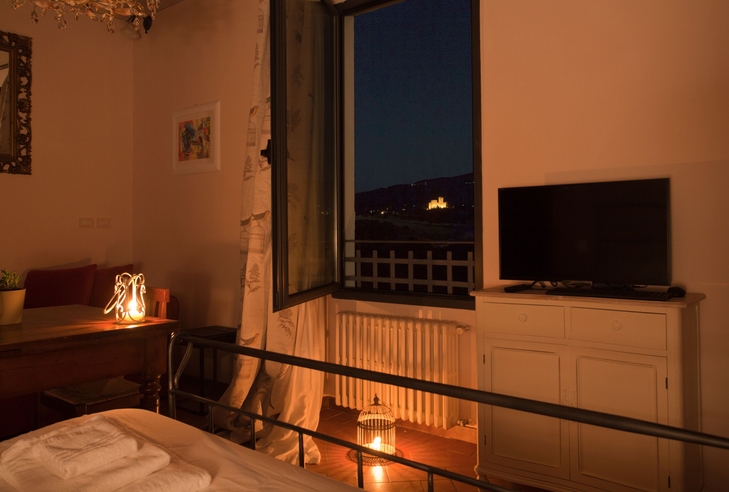 Un dormitorio con cama, una mesita de noche con lámpara, una ventana con vista a un edificio y un televisor sobre un mueble.