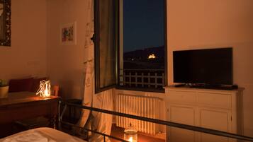 Un dormitorio con cama, una mesita de noche con lámpara, una ventana con vista a un edificio y un televisor sobre un mueble.