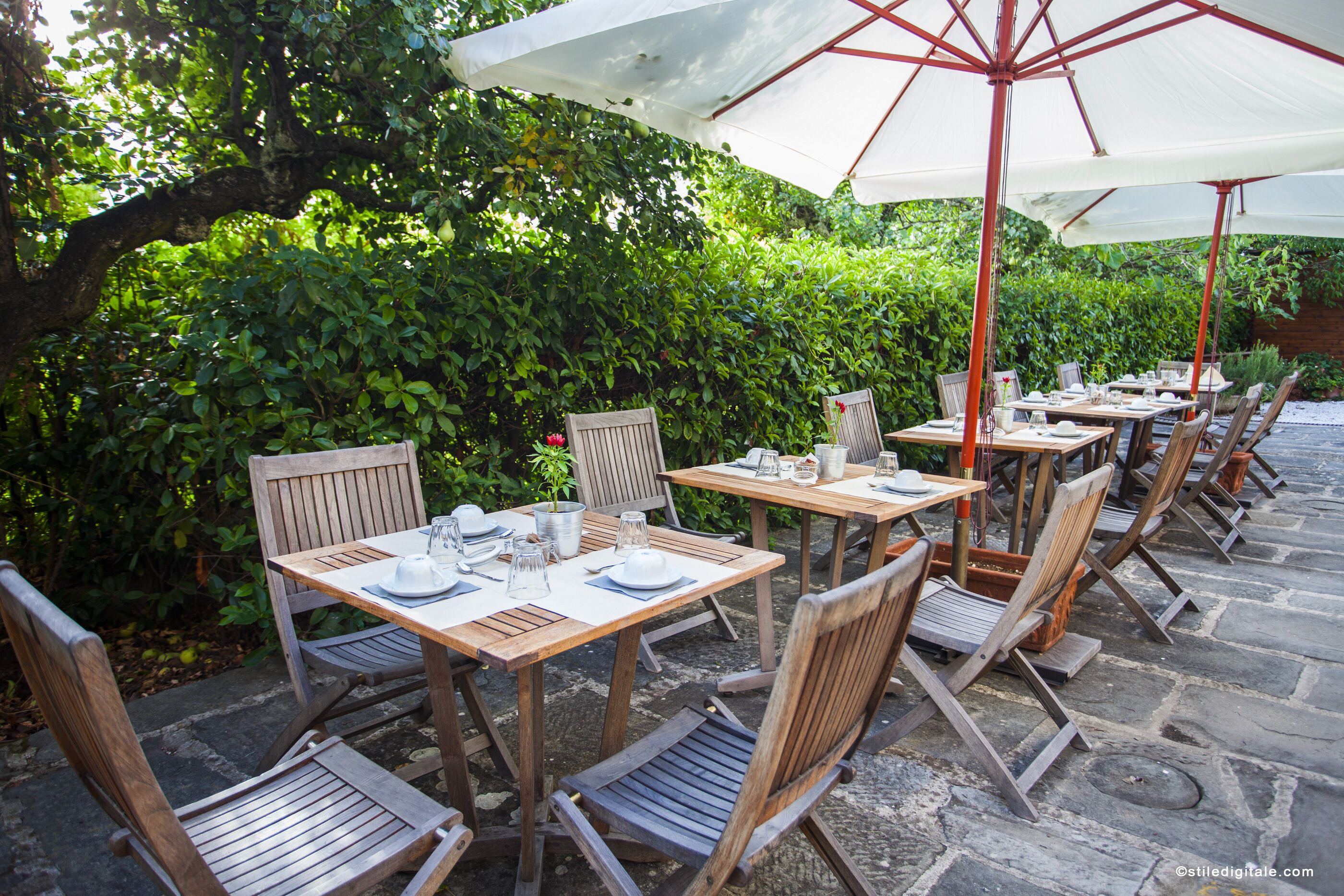 An outdoor patio with wooden tables and chairs, some under umbrellas, surrounded by lush greenery.
