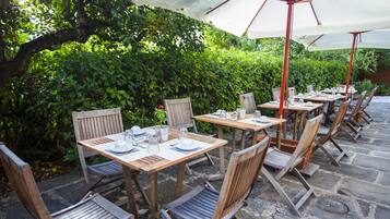 An outdoor patio with wooden tables and chairs, some under umbrellas, surrounded by lush greenery.