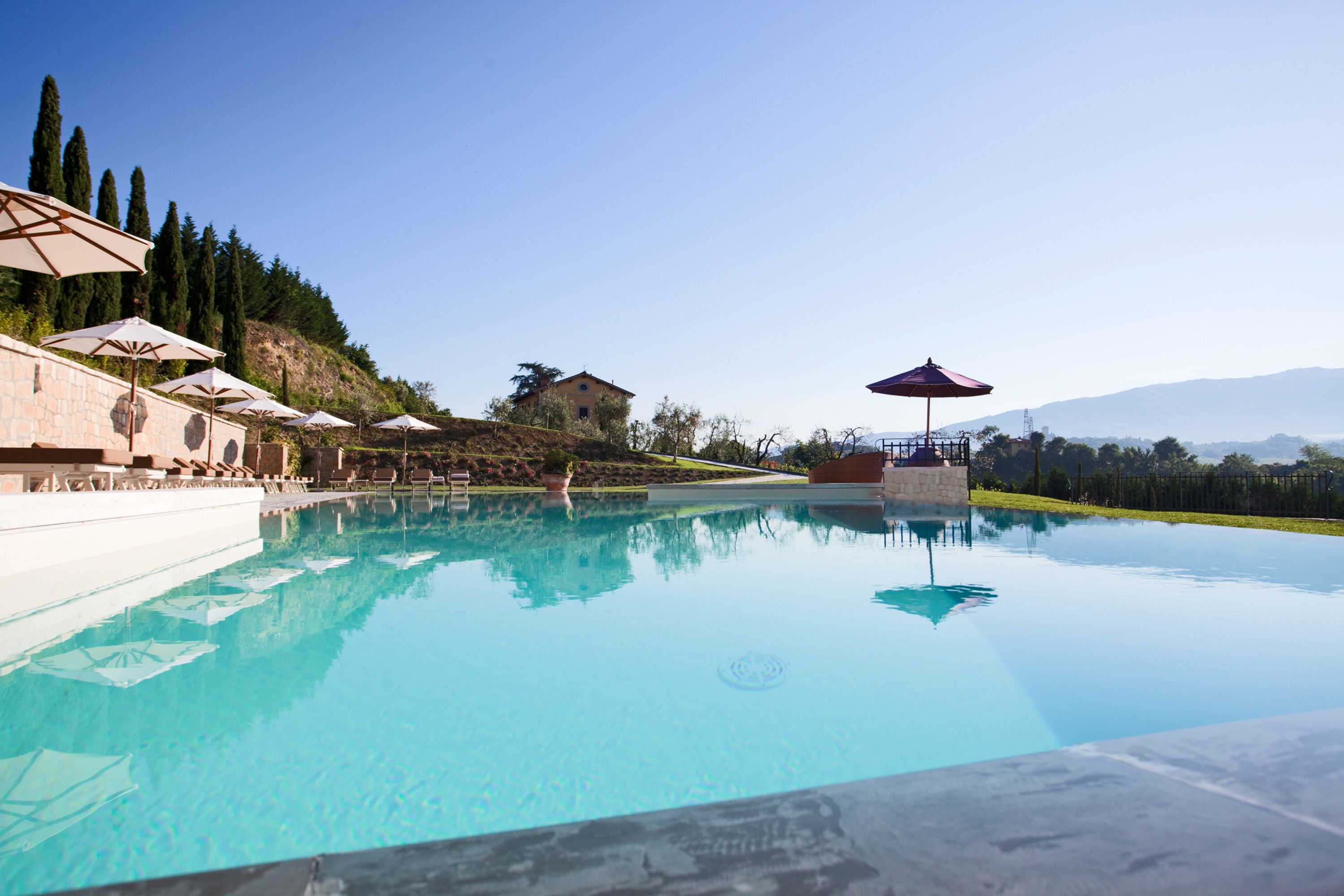 Ein klares Schwimmbecken mit Liegestühlen und Sonnenschirmen, umgeben von einer angelegten Grünfläche mit Bäumen und einer Hügelkulisse im Hintergrund.