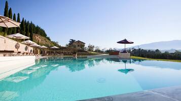 A clear swimming pool with lounge chairs and umbrellas, surrounded by a landscaped area with trees and a hill in the background.