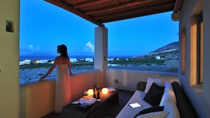 A balcony with a view of the beach and distant buildings, a wicker sofa, a small table with a bottle and glasses, and a person in a dress.
