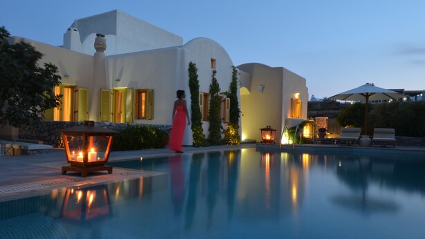 A pool area with a clear view of the pool, a person in a pink dress, and a building with yellow shutters and a white facade.