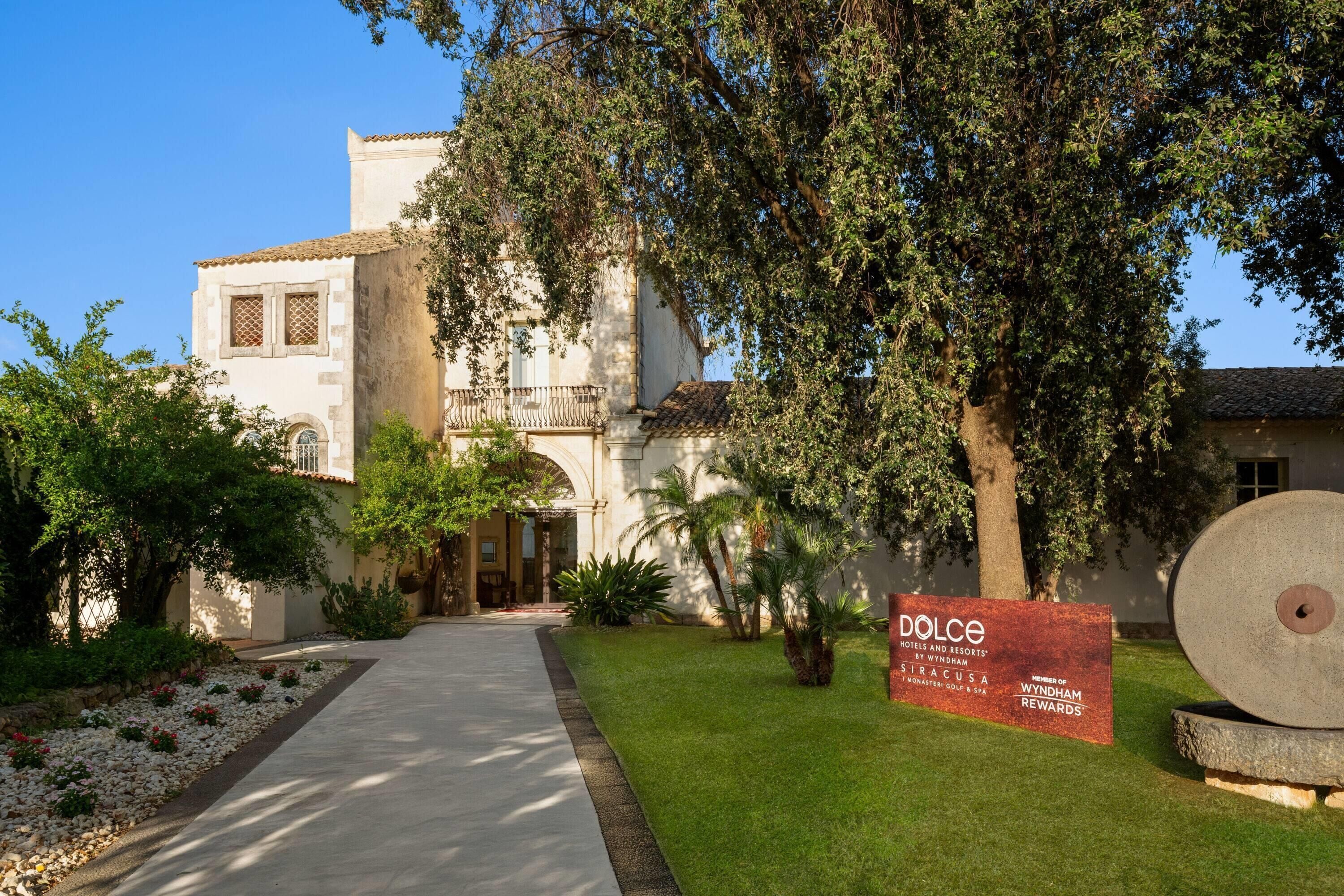 A building with a paved pathway leading to the entrance, surrounded by greenery and a sign indicating it's part of the Dolce Hotels and Resorts.