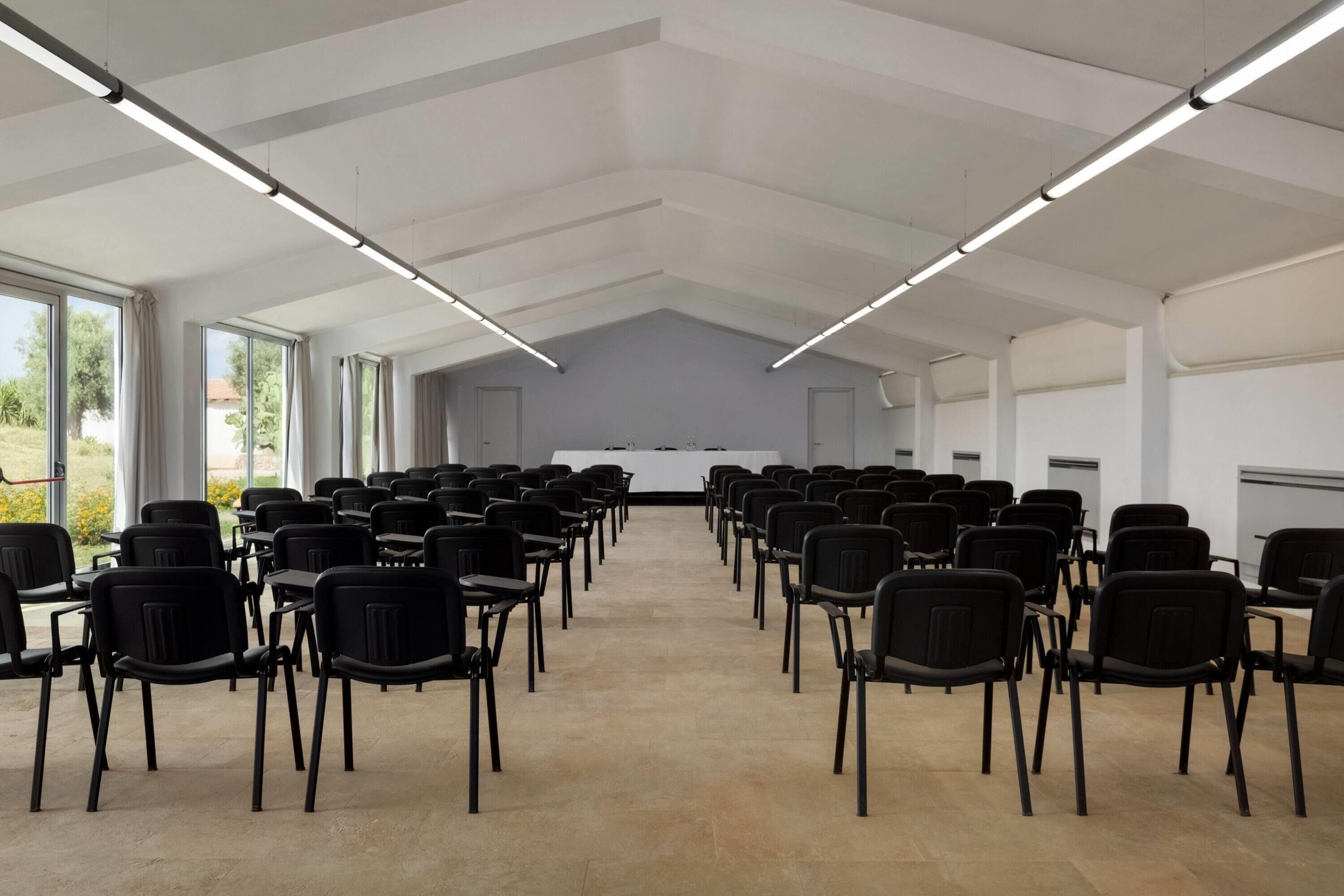 A conference room with rows of black chairs facing a white table, large windows on one side, and a high ceiling with linear lighting fixtures.