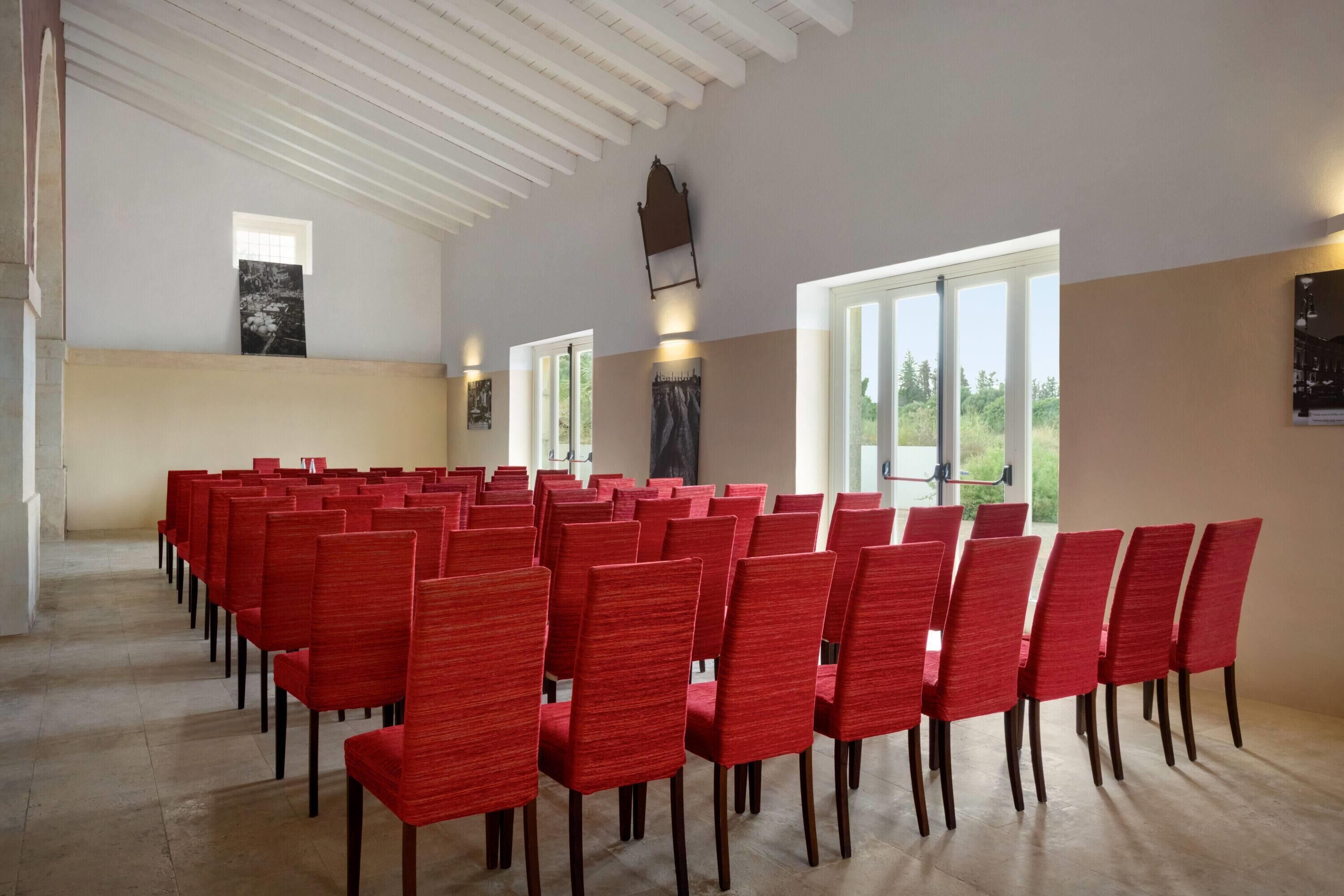 Un salón de conferencias con hileras de sillas rojas, ventanas amplias y un techo alto con vigas de madera.