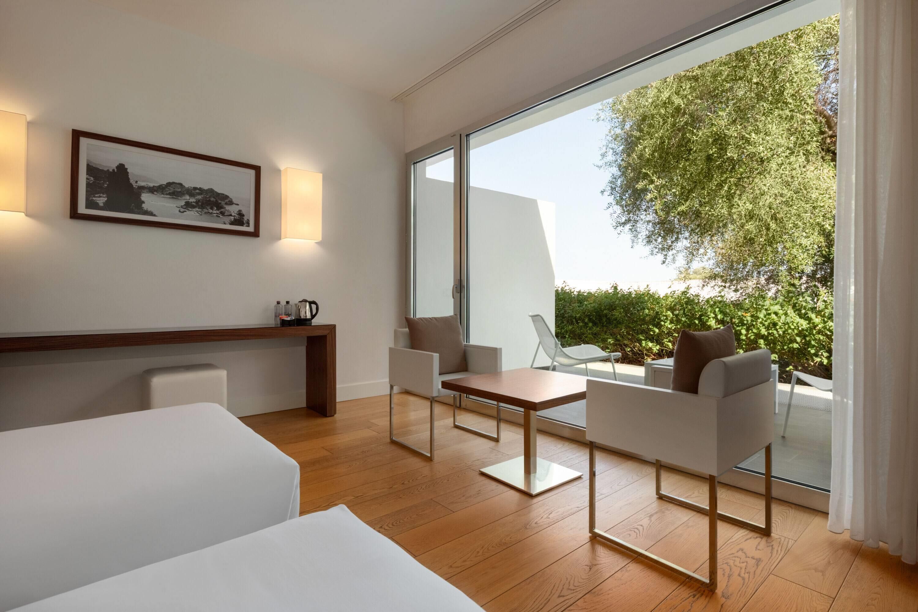 Ein modernes Hotelzimmer mit einer großen Schiebeglasdoor zum Balkon, Holzboden, einem Bett, einem Schreibtisch und zwei Stühlen.