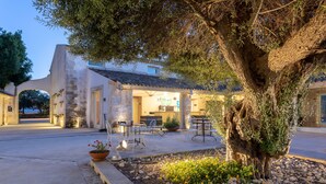 An outdoor seating area with tables and chairs, a large tree, and a building with a tiled roof. - Dolce by Wyndham Siracusa I Monasteri Golf & Spa (Syracuse)