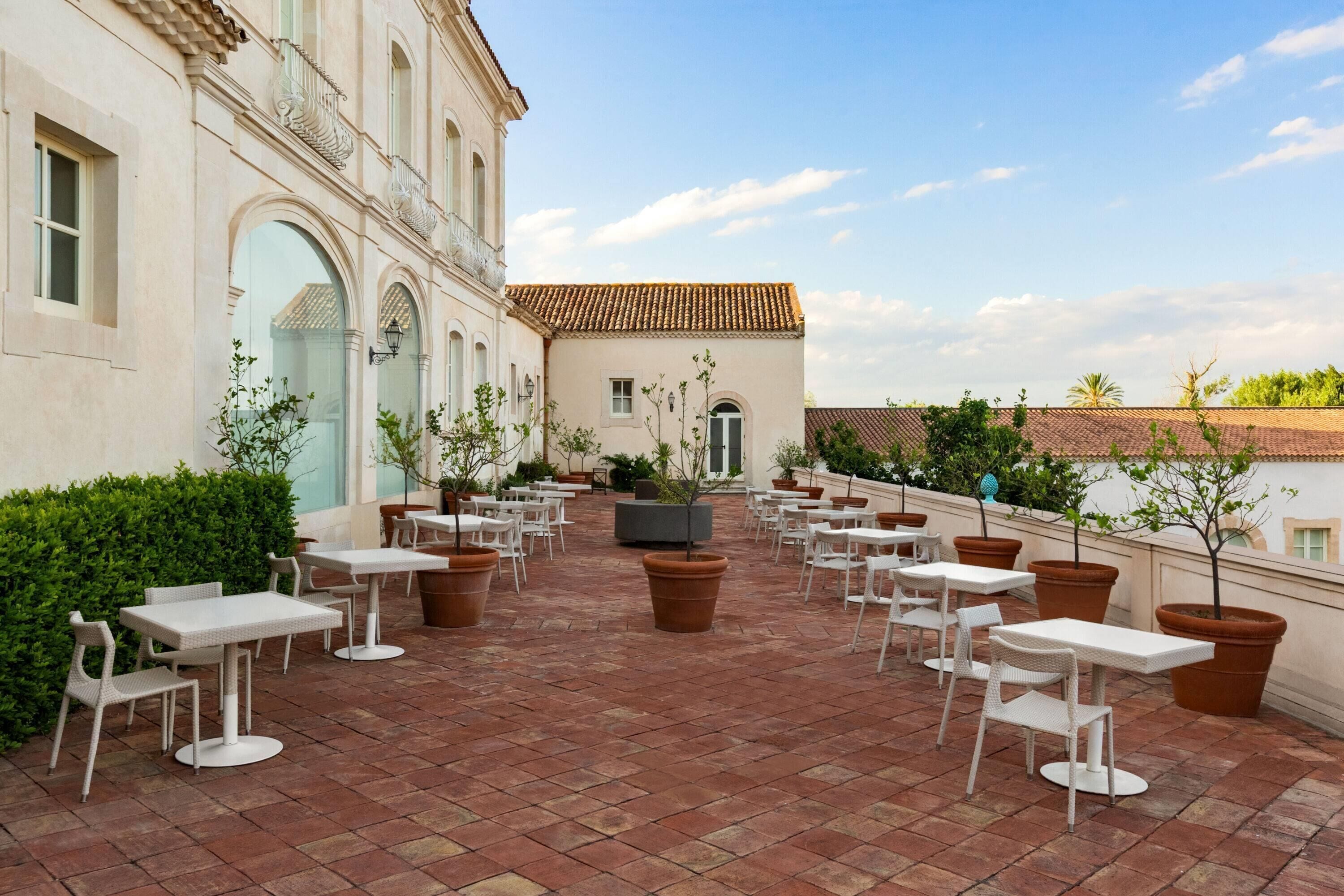 Un patio exterior con mesas y sillas blancas, rodeado de plantas en macetas y un edificio con tejado de baldosas.